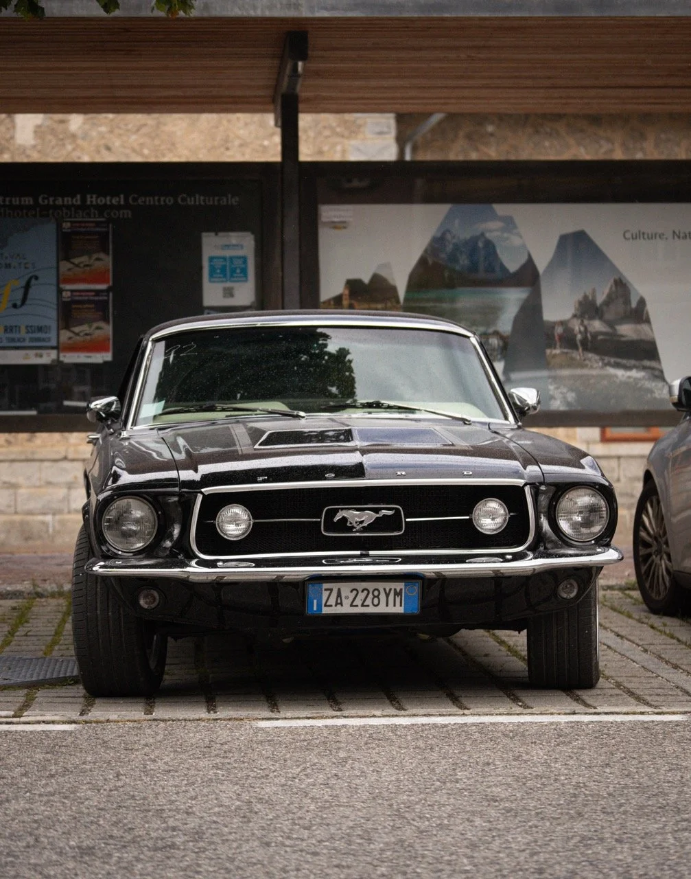 L'immagine mostra una Ford Mustang d'epoca nera parcheggiata di fronte a un edificio con poster e fotografie di paesaggi.