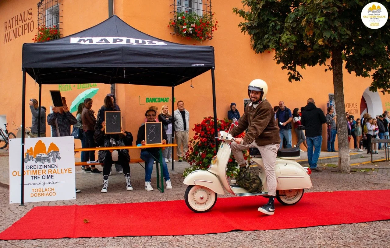 Uomo che sorride su una vespa bianca con casco, sul red carpet davanti a un edificio arancione, con persone in fila e uno stand con cartelli e un banco in una piazza