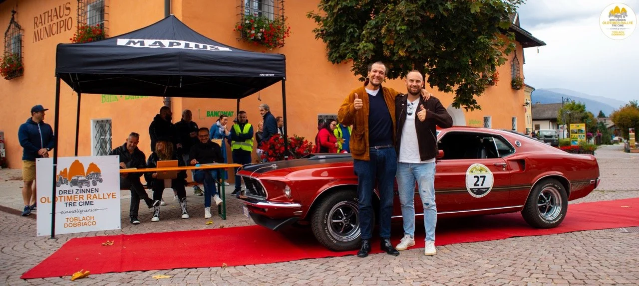 Due uomini felici davanti a una macchina d'epoca rossa alla rievocazione di rally, con un edificio arancione di sfondo e un'area per il pubblico.
