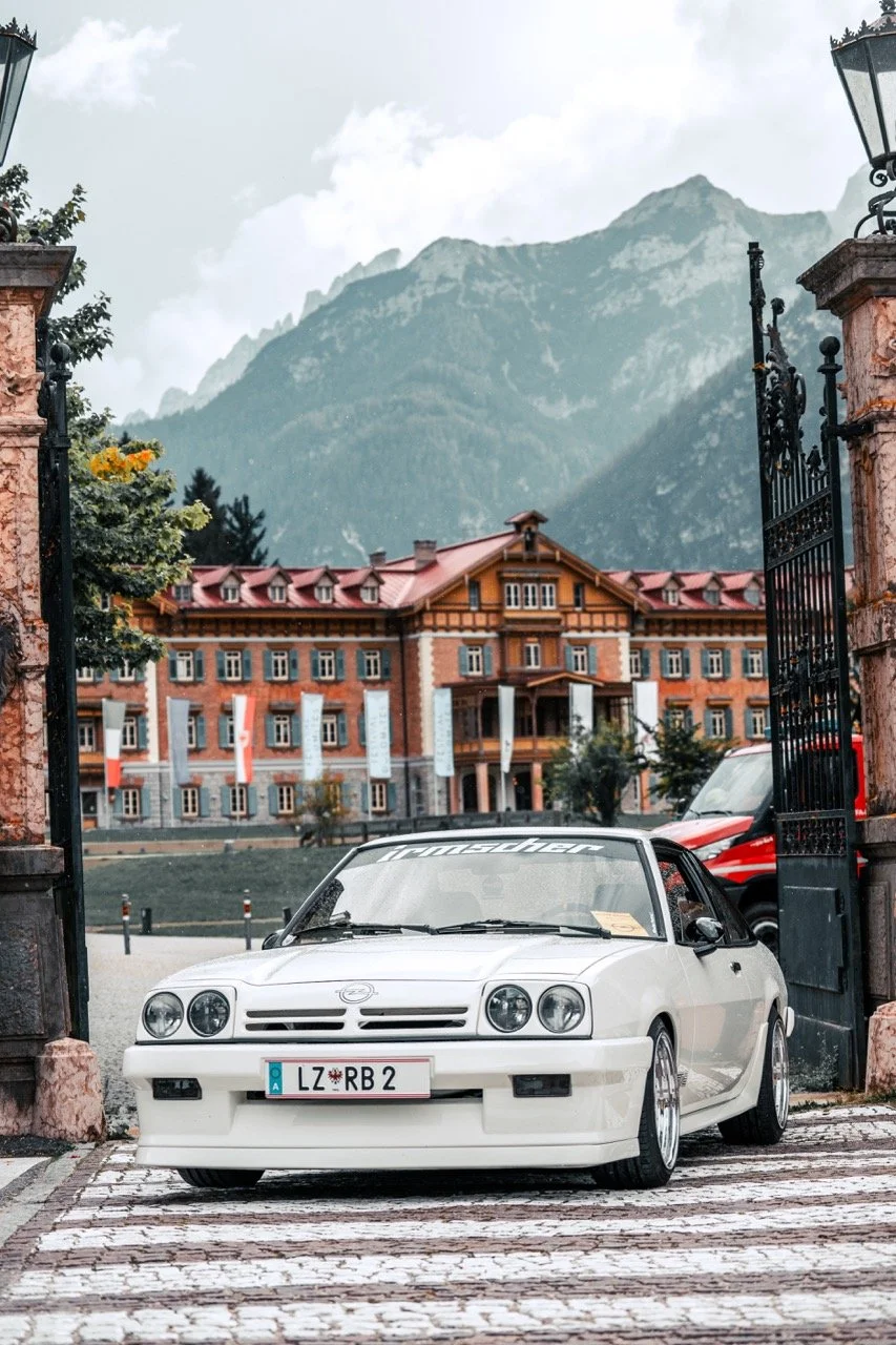 Auto sportiva bianca parcheggiata davanti a un grande edificio in stile alpino con montagne sullo sfondo.