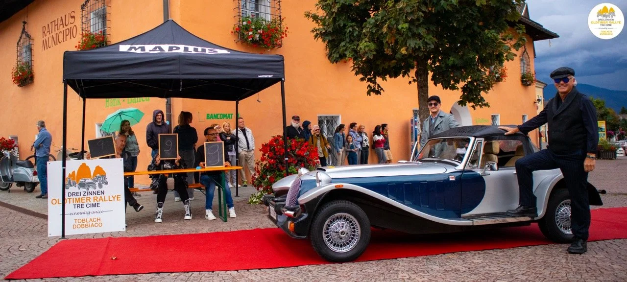 Auto d'epoca con due uomini in abiti vintage e un'auto d'epoca parcheggiata su un tappeto rosso davanti a un edificio storico con persone in fila e in piedi vicino a un banco di registrazione per un rally d'epoca in Italia.