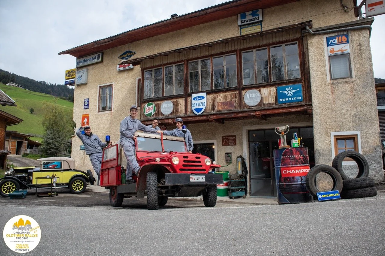 Gruppo di persone in abbigliamento da officina meccanica vicino a un veicolo rosso e un'altra auto d'epoca in un'officina di montagna con vari pneumatici e attrezzi, su una strada di montagna.