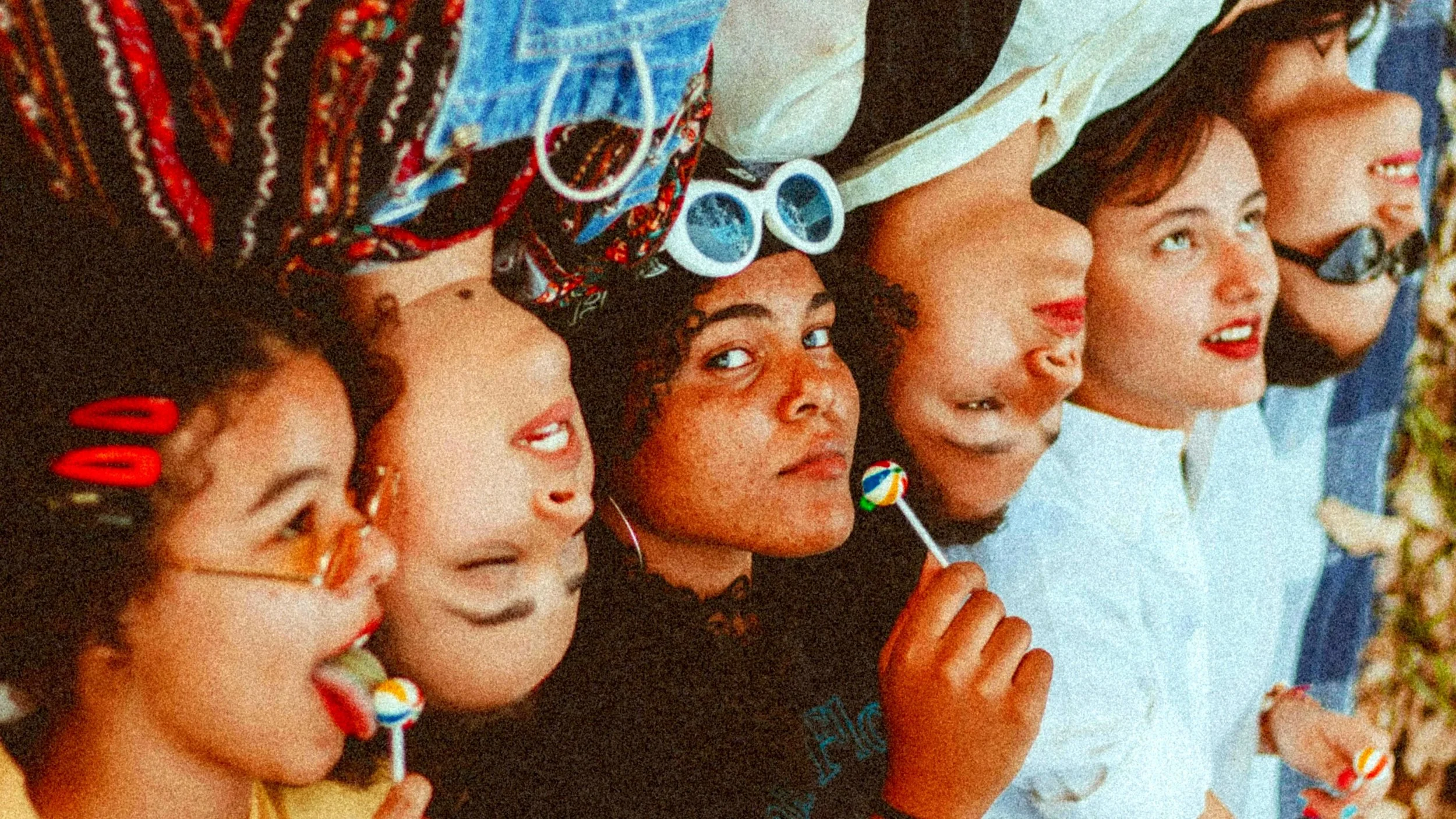 A group of six young women lying on their backs, head close together, smiling and holding colorful lollipops, with some wearing glasses or sunglasses, in a festive and playful setting.