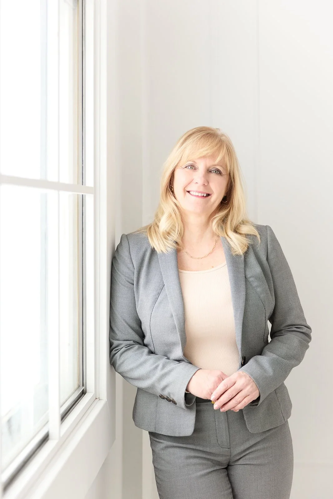 A woman with blonde hair wearing a gray suit standing by a window and smiling.