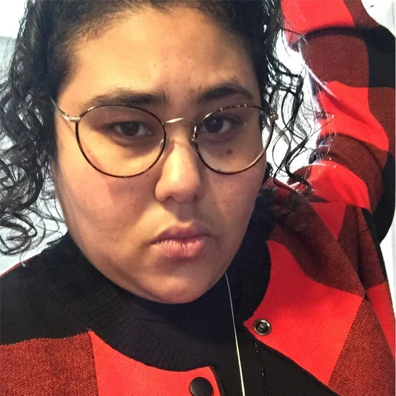 Close-up of a person with short curly hair and round glasses, wearing a red and black checkered jacket, looking directly at the camera.