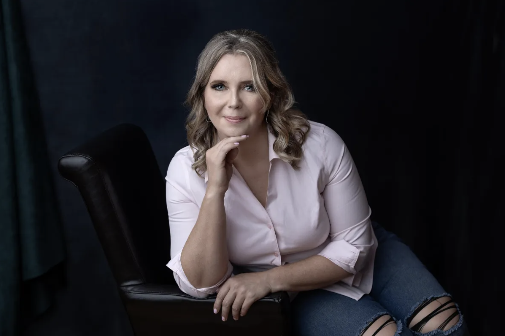 A woman with blonde hair, wearing a light pink shirt and ripped jeans, sitting on a dark chair with a dark background, smiling at the camera.