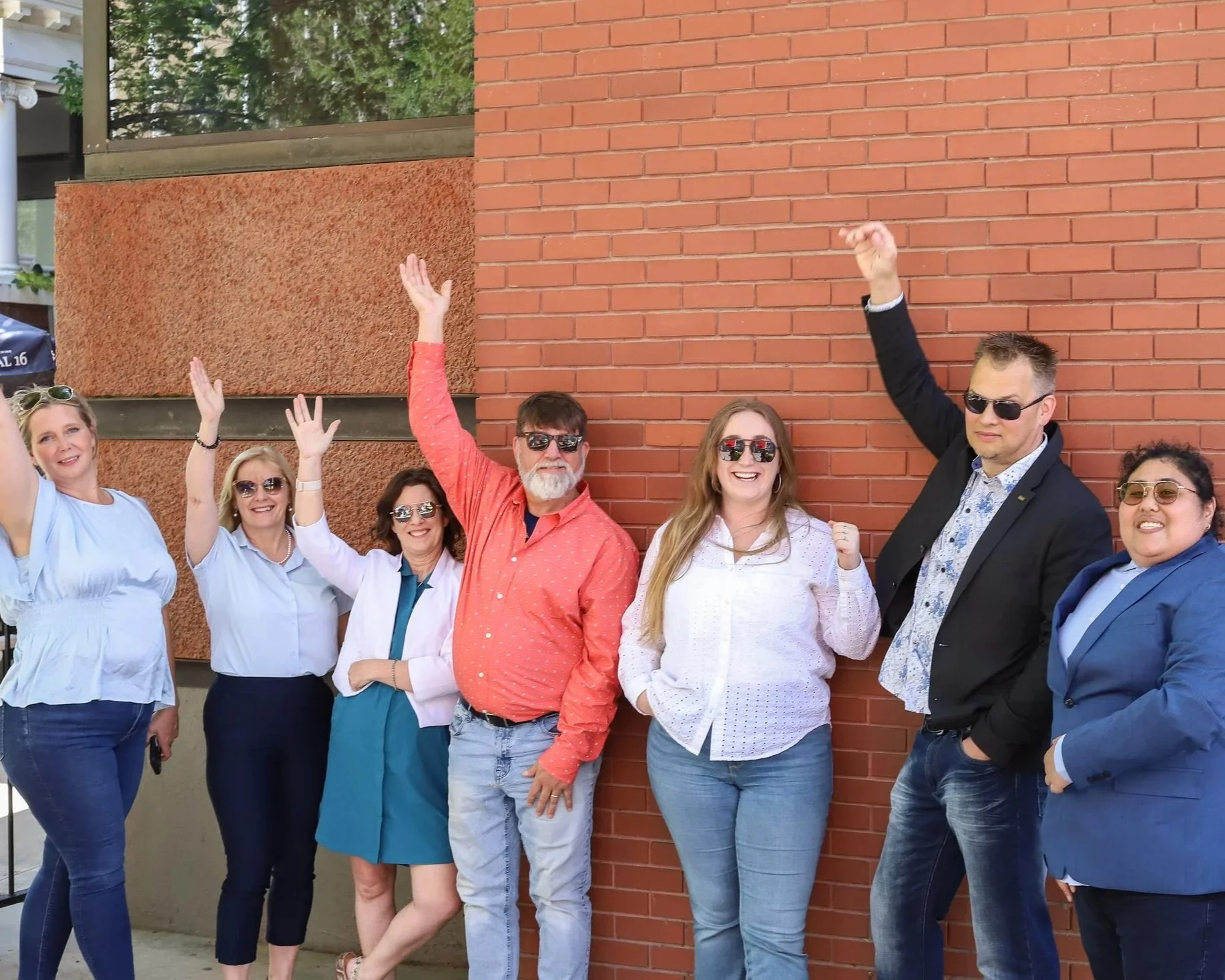 A diverse group of seven people standing against a brick wall, smiling, raising their hands and fists in celebration, wearing sunglasses, dressed in casual and business attire.