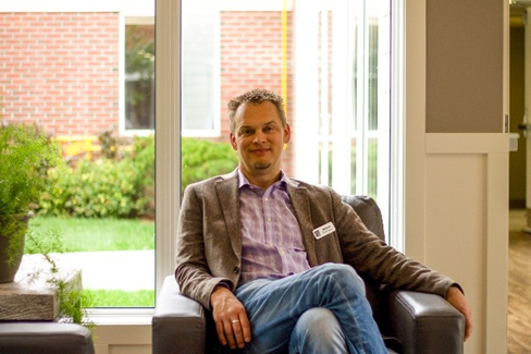 A man sitting in a black leather chair in a well-lit room with large windows showing a garden outside.