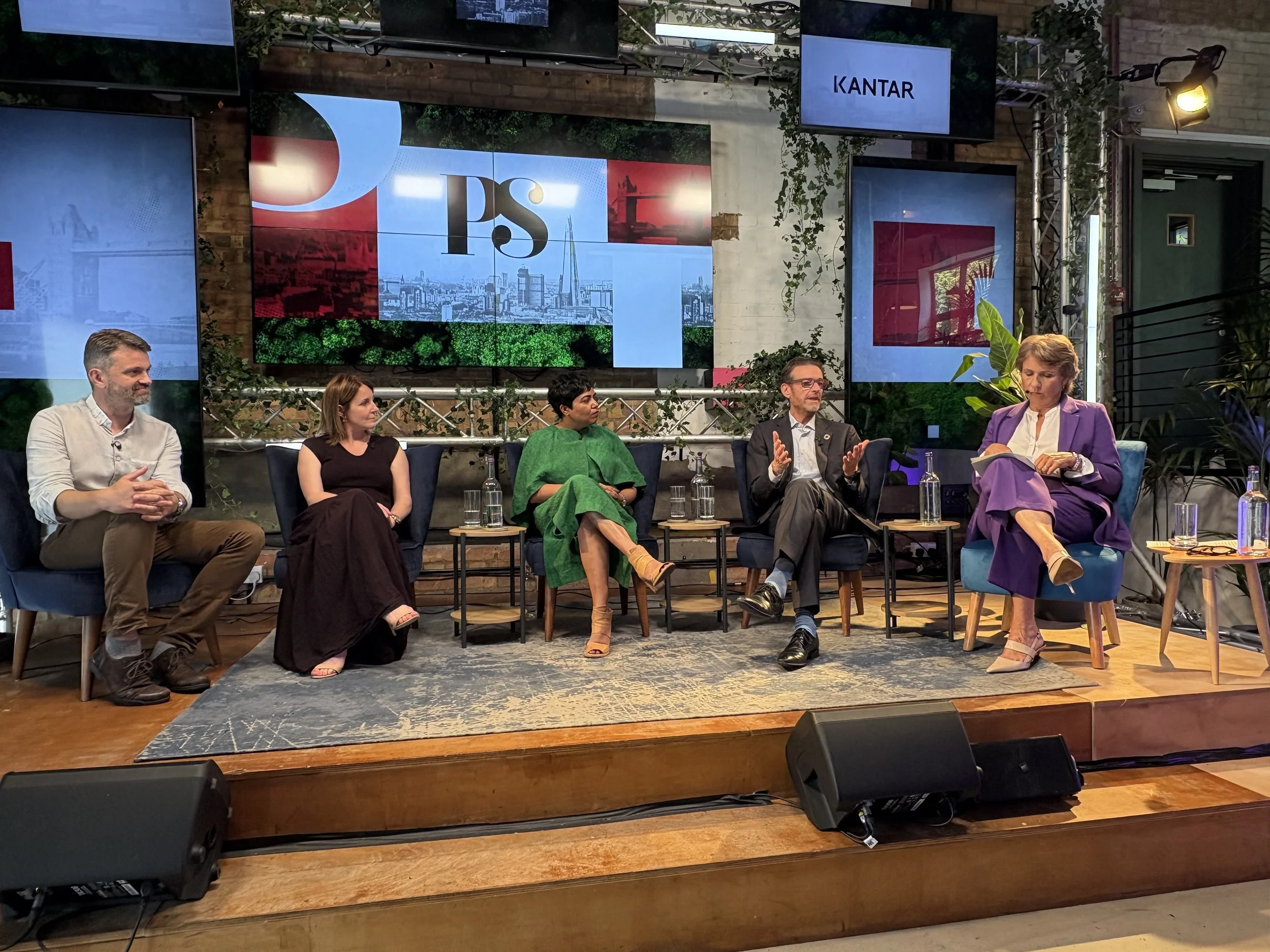 Preeti Srivastav participating in a panel discussion, with multiple large screens displaying graphics behind them, set in a venue with plants and industrial decor.