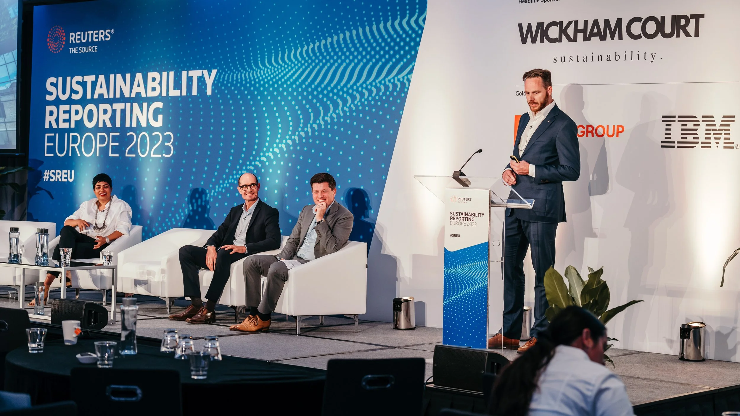 A panel discussion at REUTERS Sustainability Reporting Europe 2023 with four people, three seated on a stage and one standing at a podium, during a conference on sustainability in Europe.
