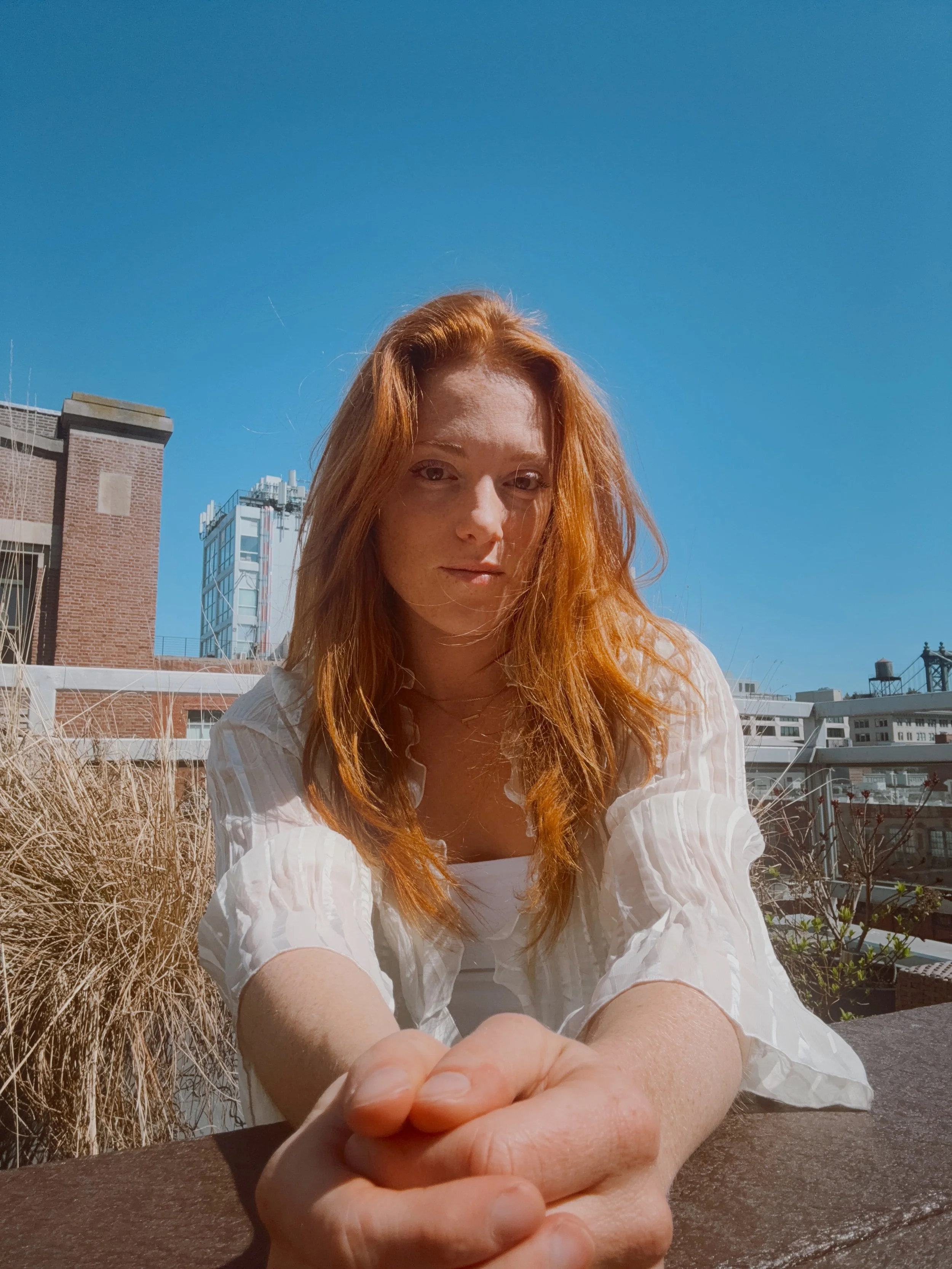 A woman with red hair sitting outdoors on a bright sunny day, leaning forward with hands clasped on a wooden surface, with buildings and a clear blue sky in the background.