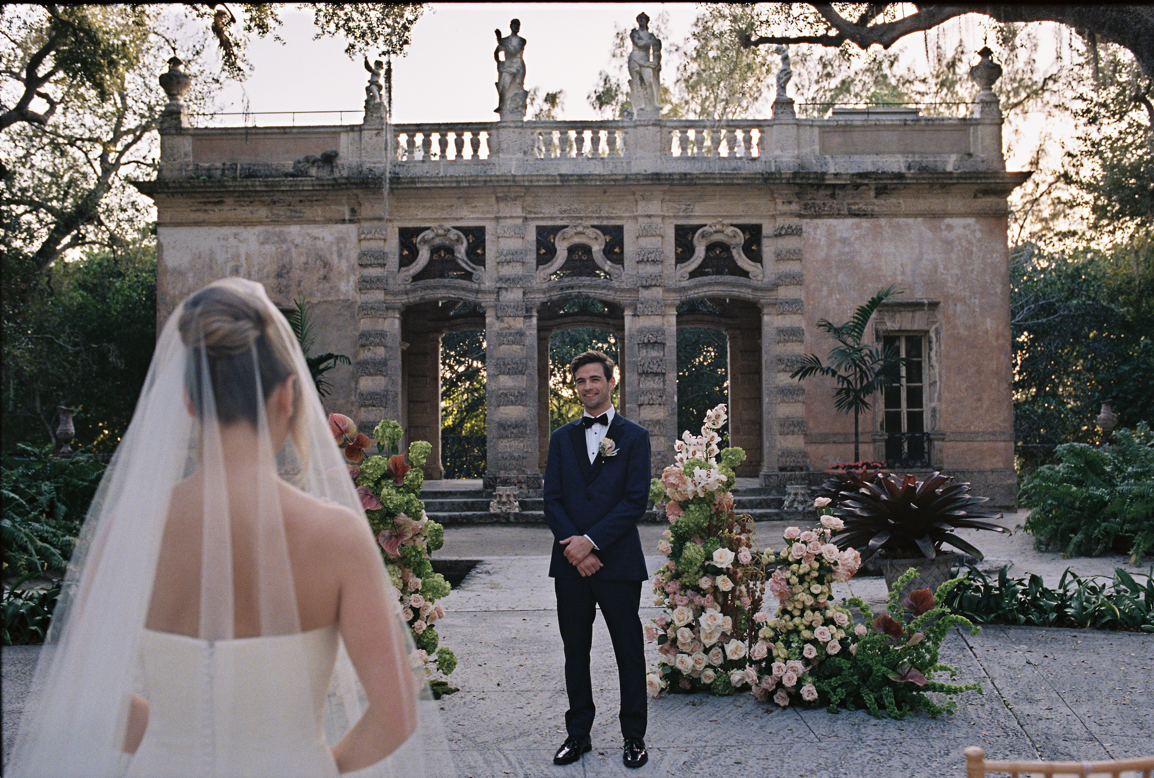 Groom waiting at the ceremony space surrounded by floral arrangements as the bride approaches during their Vizcaya Museum and Gardens wedding.