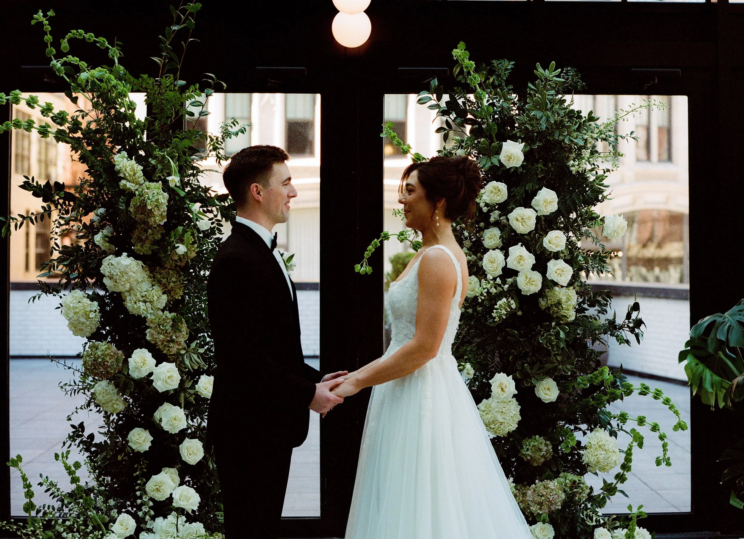 Bride and groom holding hands in front of lush white floral ceremony arrangements inside Shinola Hotel