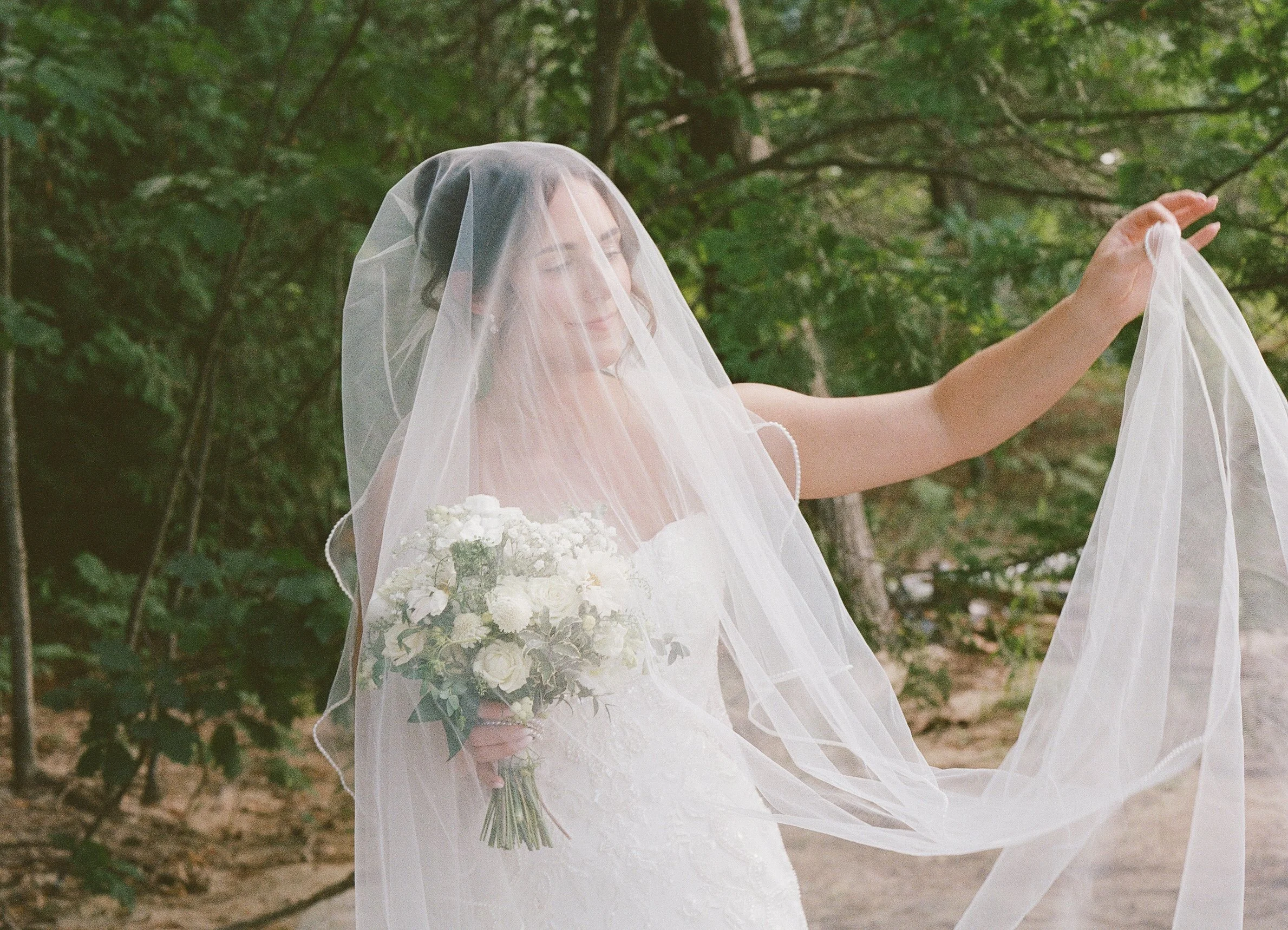Bride holding her white bouquet with her long veil draped over her face outdoors.