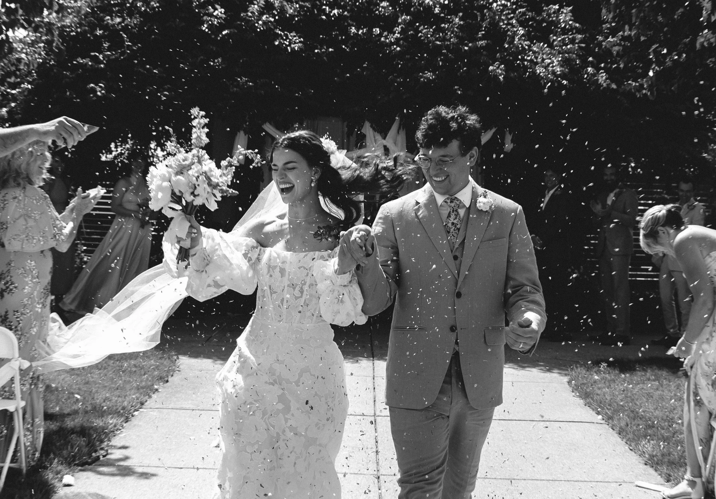 Newlyweds walking through falling flower petals, holding hands and smiling, captured in a joyful black and white film wedding photography moment