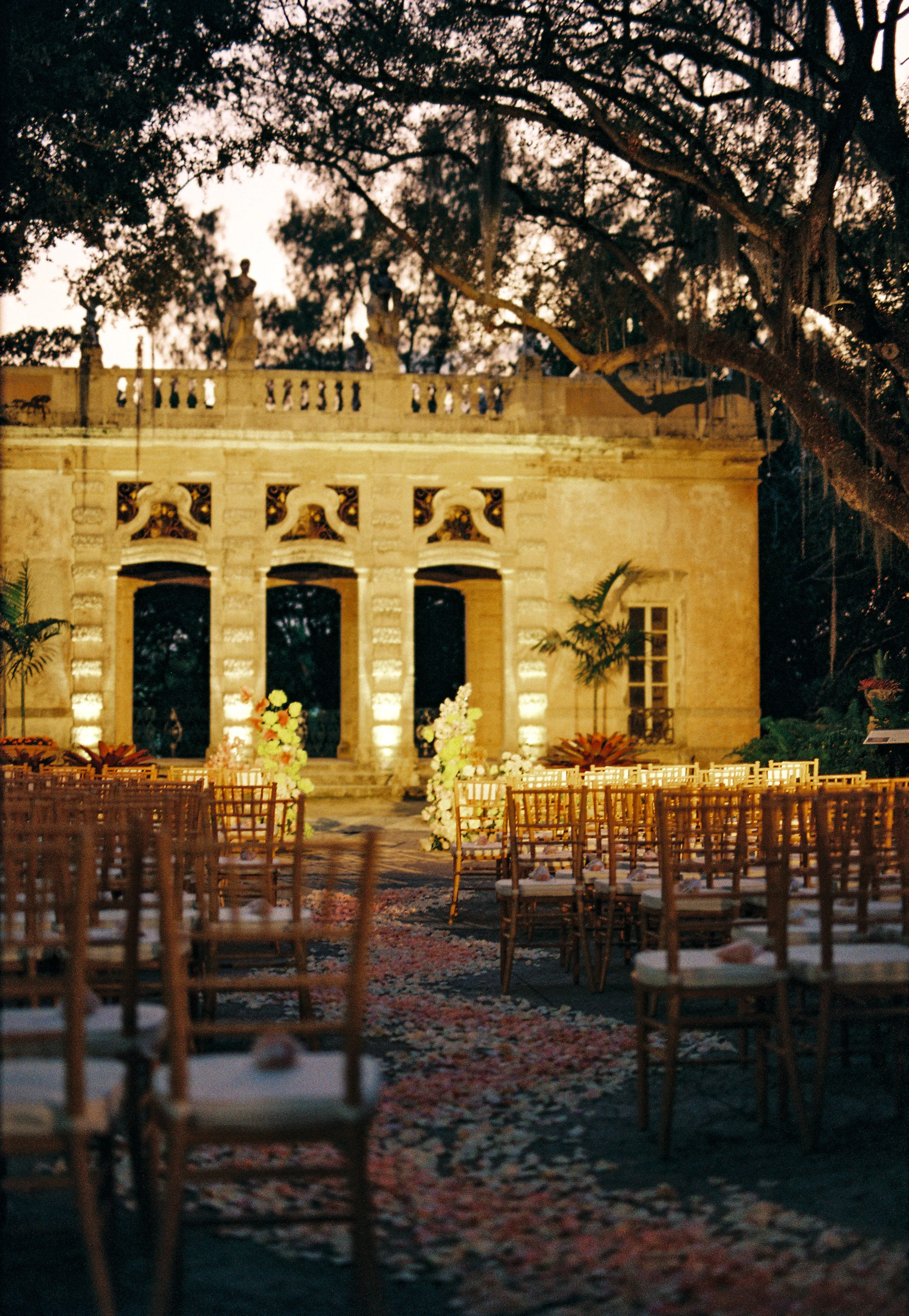 Outdoor ceremony setup at Vizcaya Museum and Gardens wedding with wooden chairs, rose petals lining the aisle, and the historic stone garden pavilion glowing in warm evening light. (Copy)