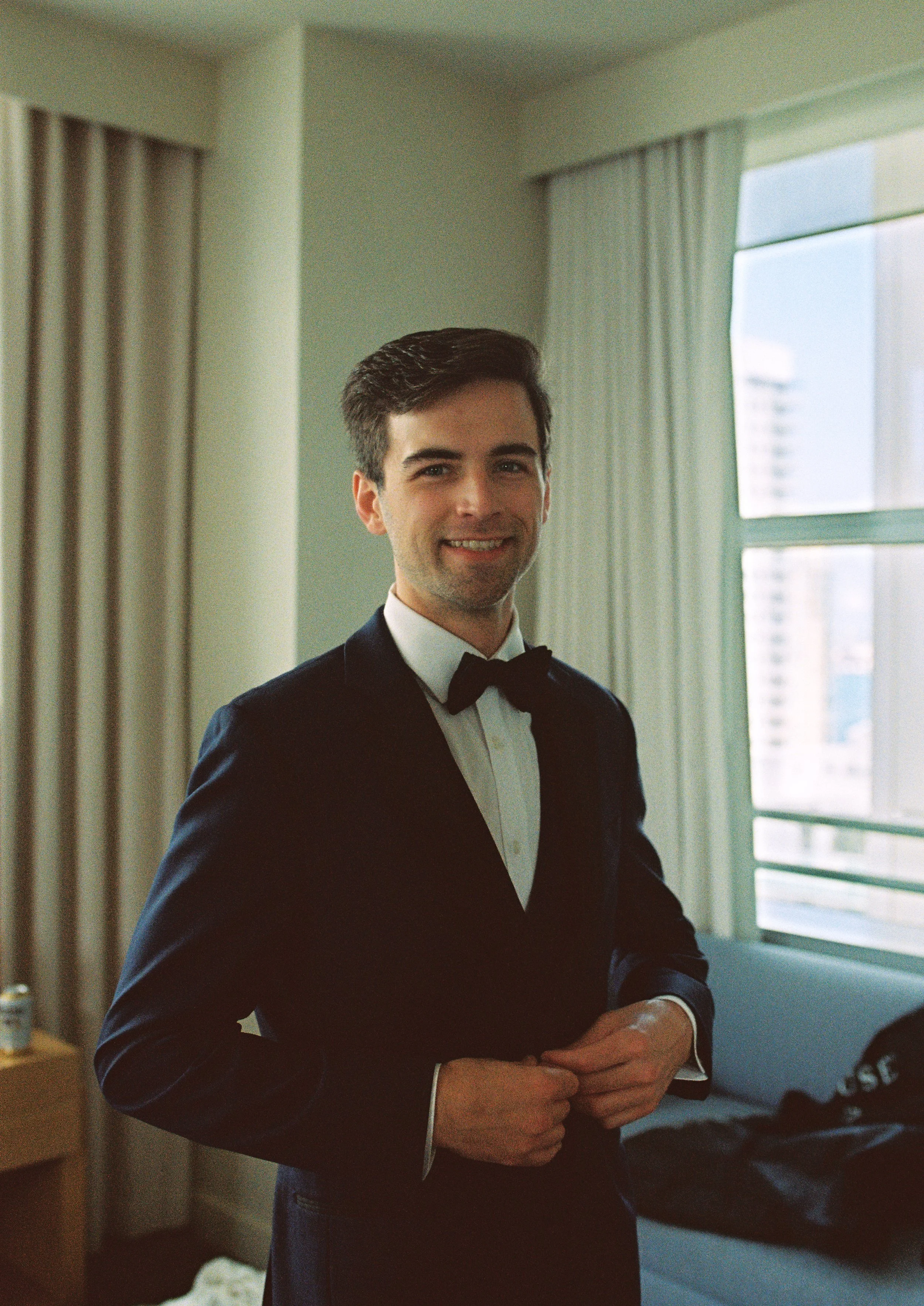 Groom in a black tuxedo adjusting his jacket while getting ready near a sunlit window. (Copy)