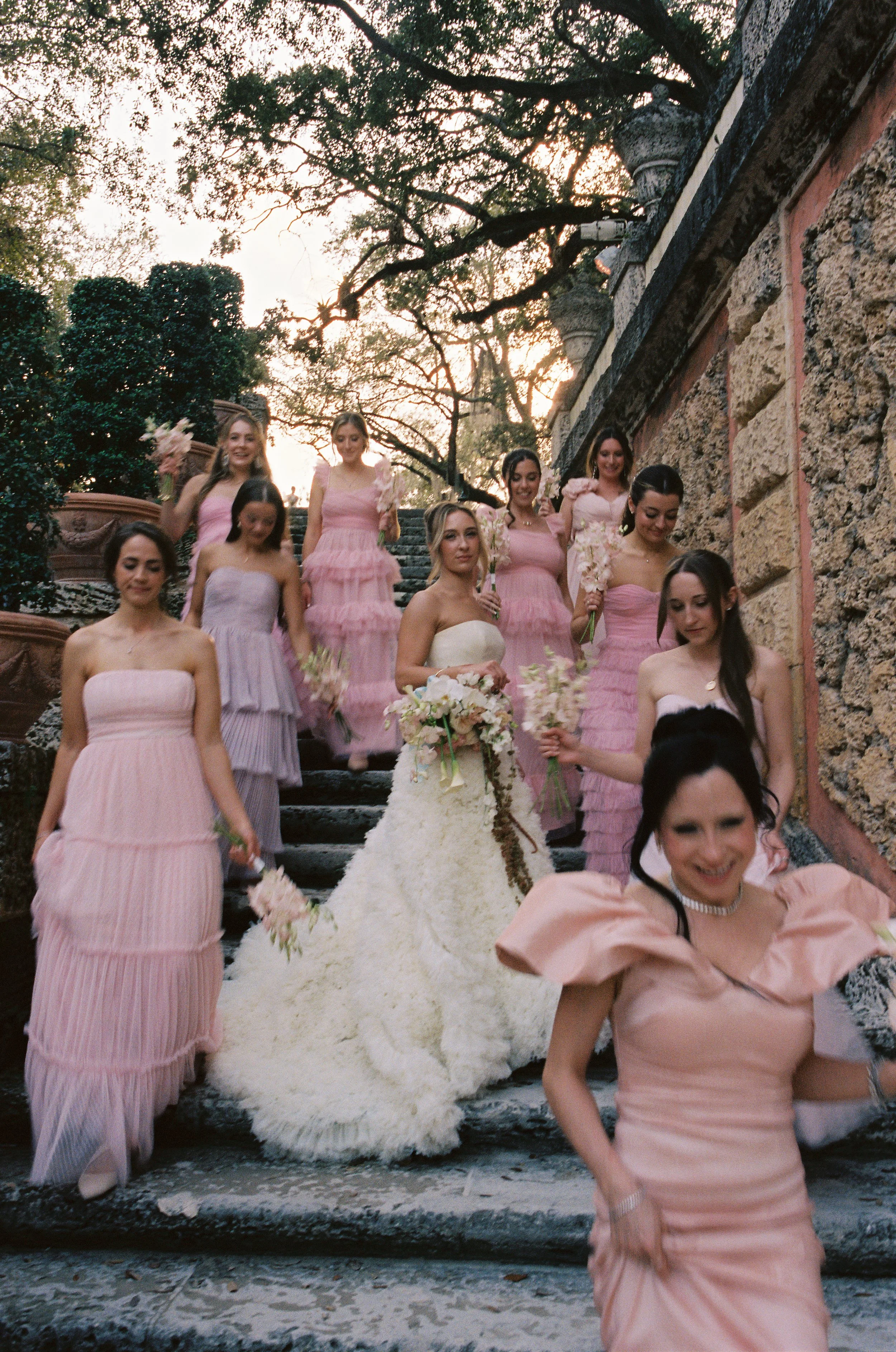Bride and bridesmaids in pink dresses walking down the historic stone staircase at Vizcaya Museum and Gardens holding bouquets. (Copy)