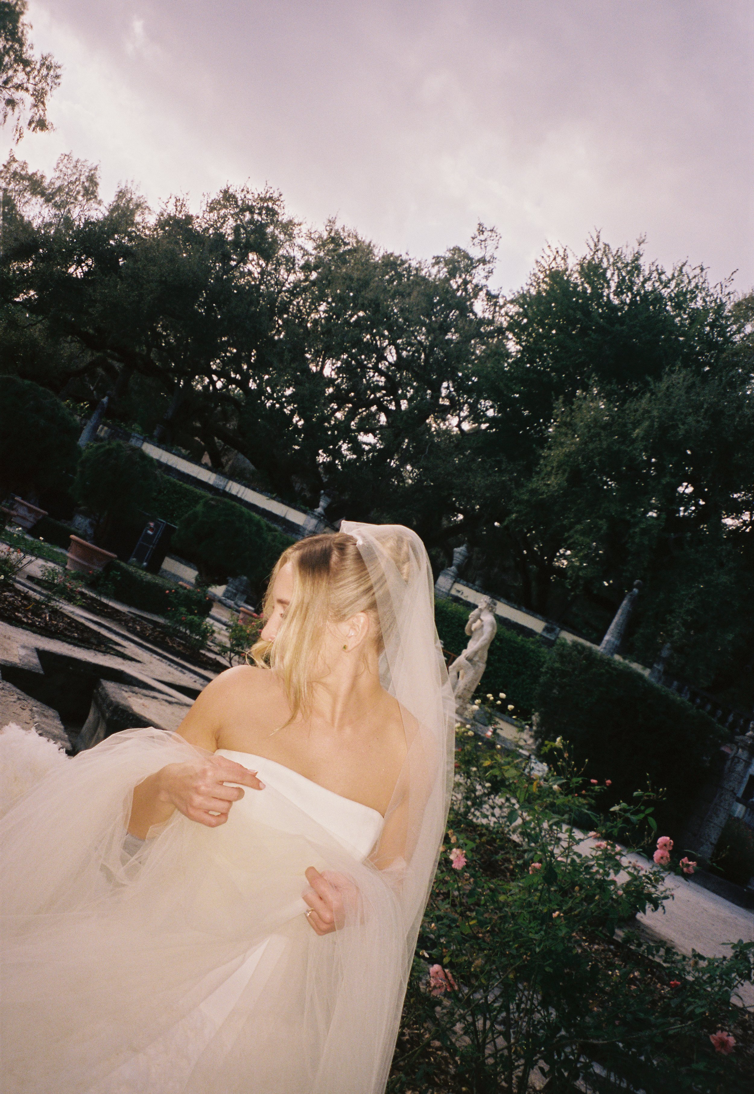 Bride in a strapless gown and flowing veil standing in the romantic gardens of Vizcaya Museum and Gardens with statues and rose bushes behind her. (Copy)