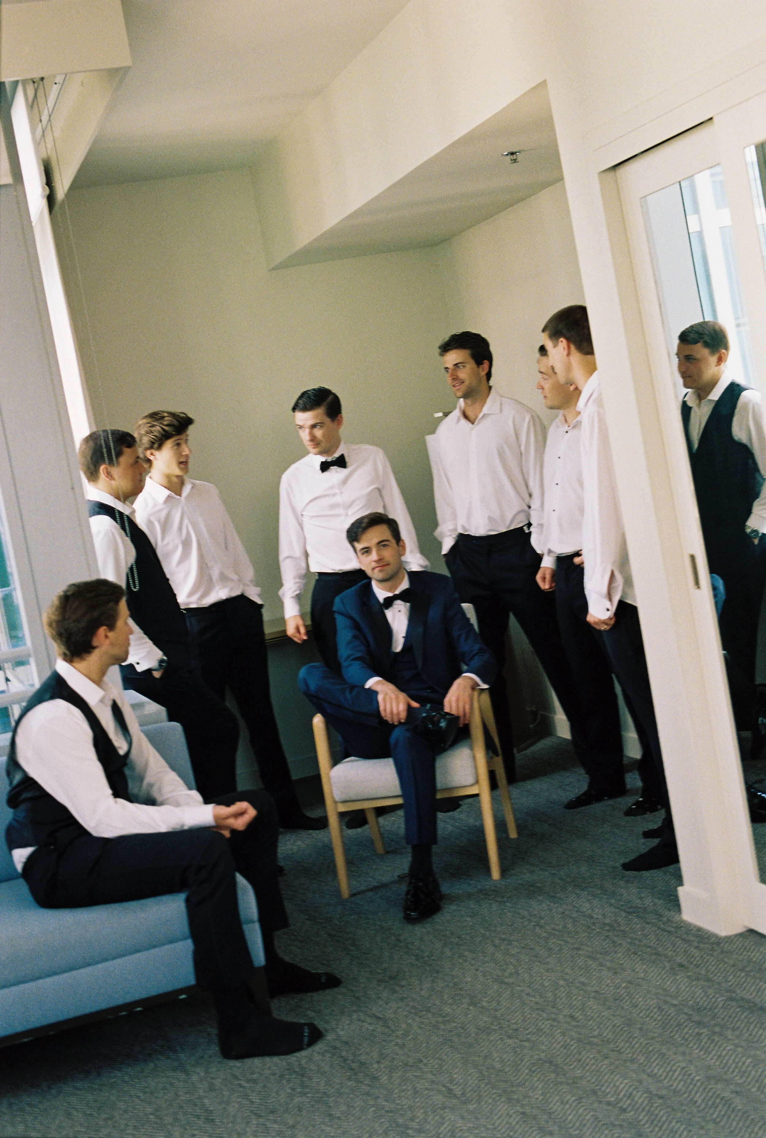 Groom getting ready with his groomsmen in a modern suite, wearing a navy tuxedo while his friends gather around before the ceremony. (Copy)
