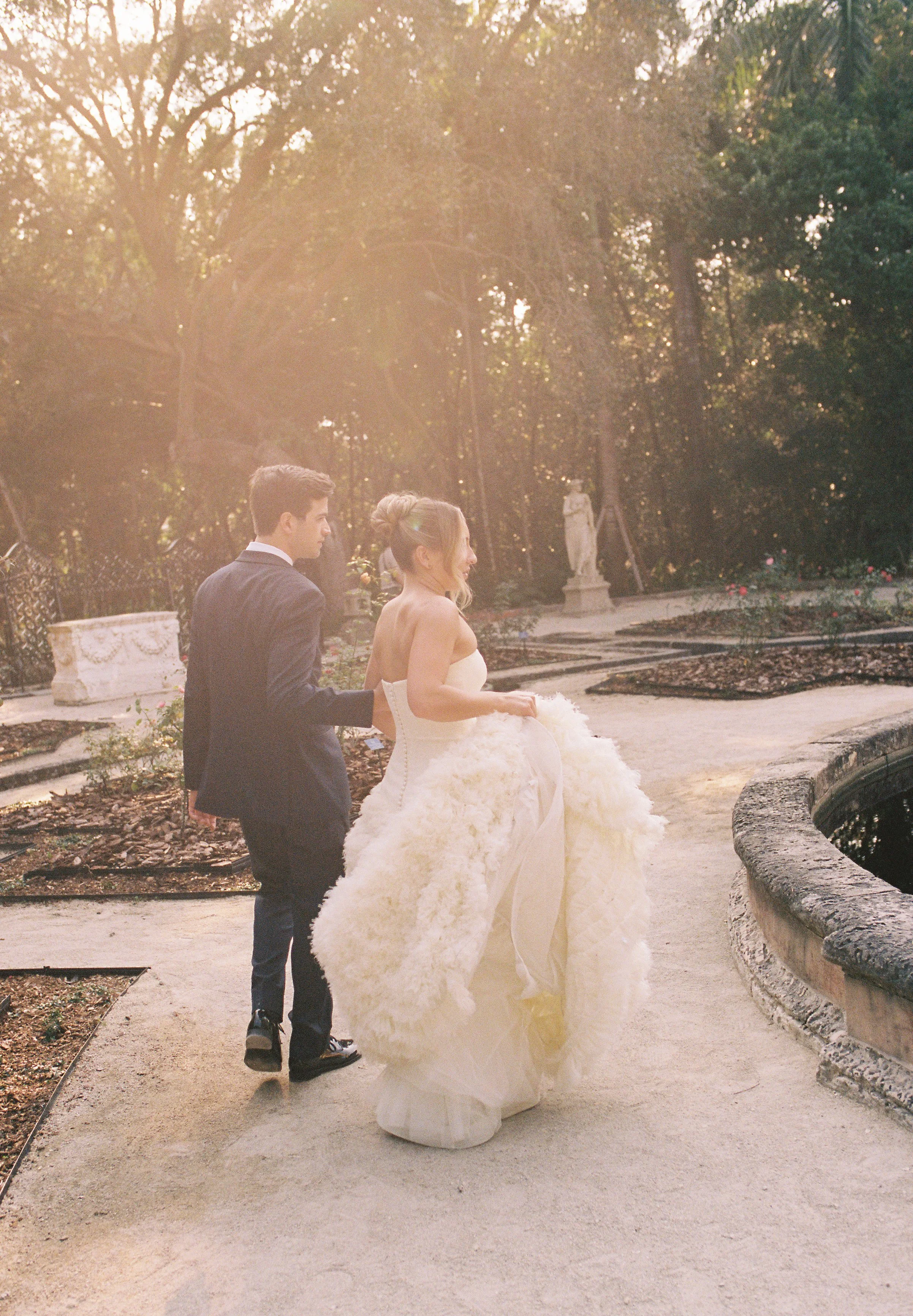 Bride and groom walking together along a garden pathway in soft golden light during their Vizcaya Museum and Gardens wedding portraits. (Copy)