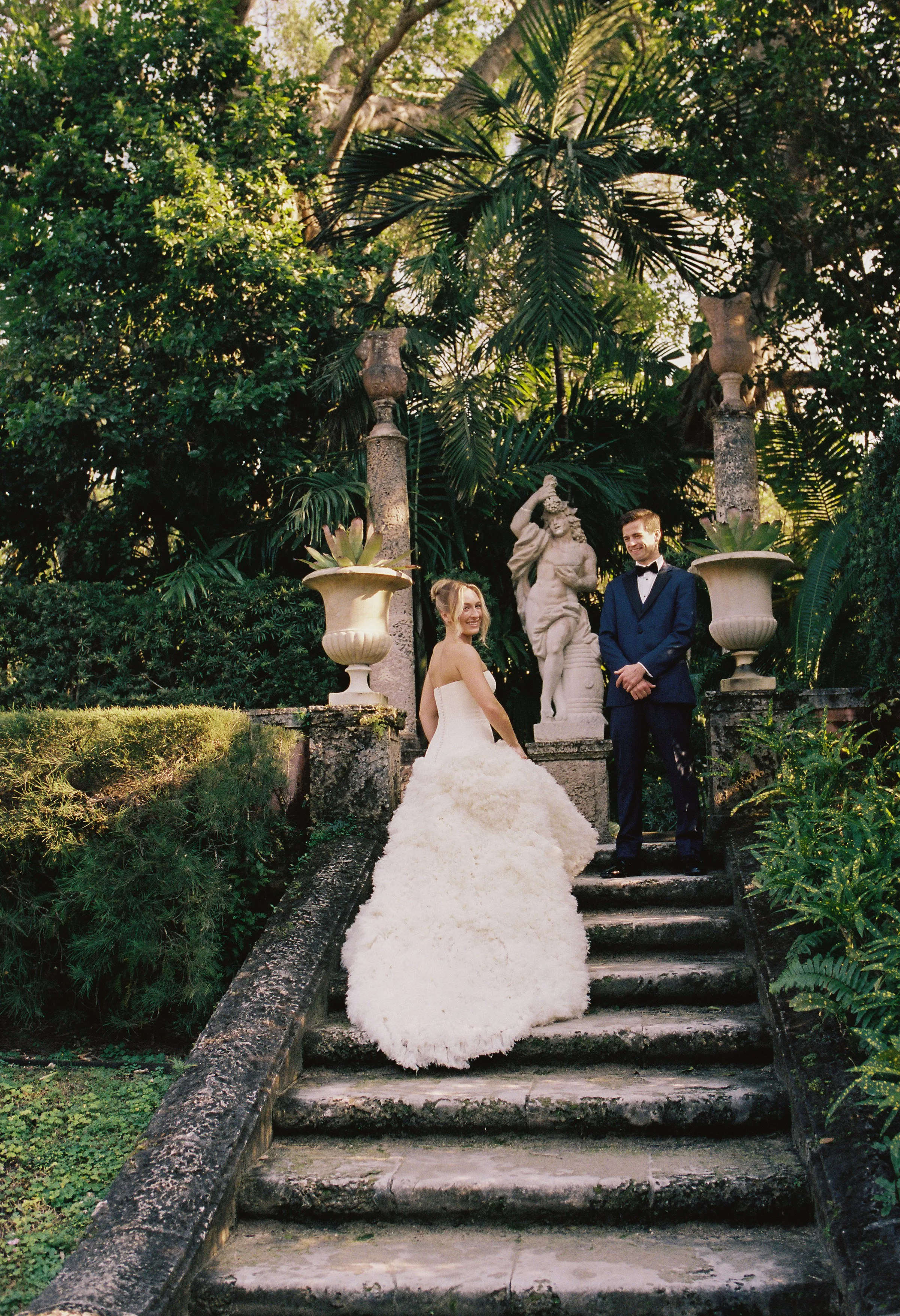 Bride and groom standing on a grand stone staircase surrounded by tropical greenery and statues during their Vizcaya Museum and Gardens wedding. (Copy)