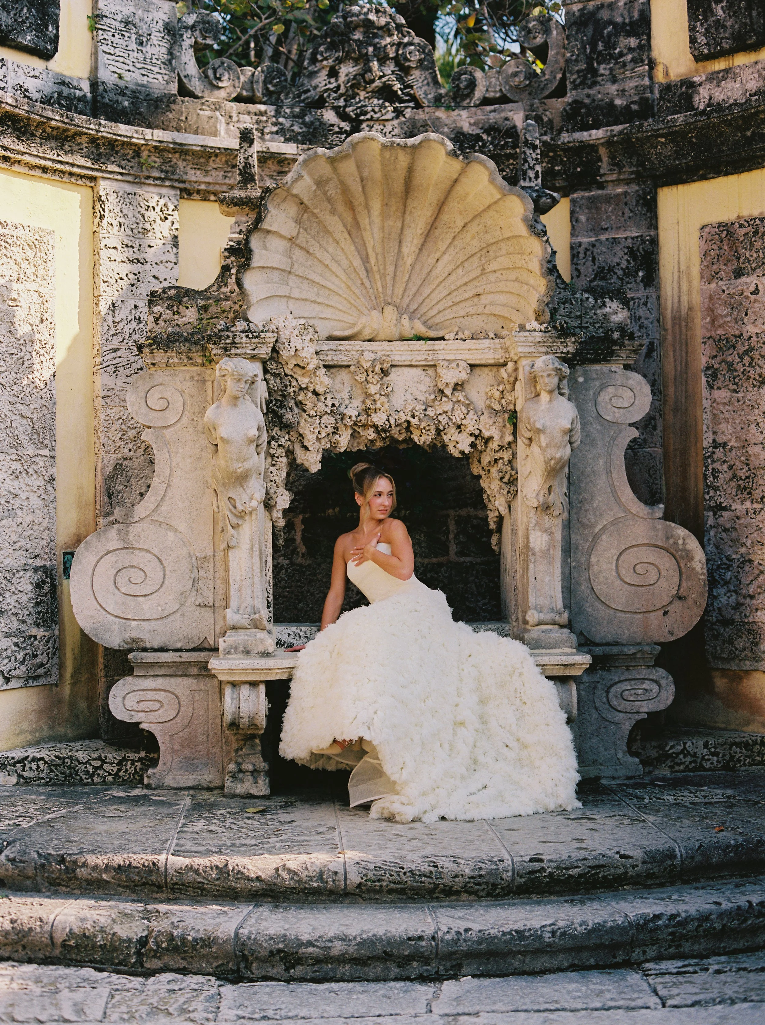 Bride sitting beneath an ornate stone shell fountain surrounded by historic sculptures in the gardens at Vizcaya Museum and Gardens. (Copy)