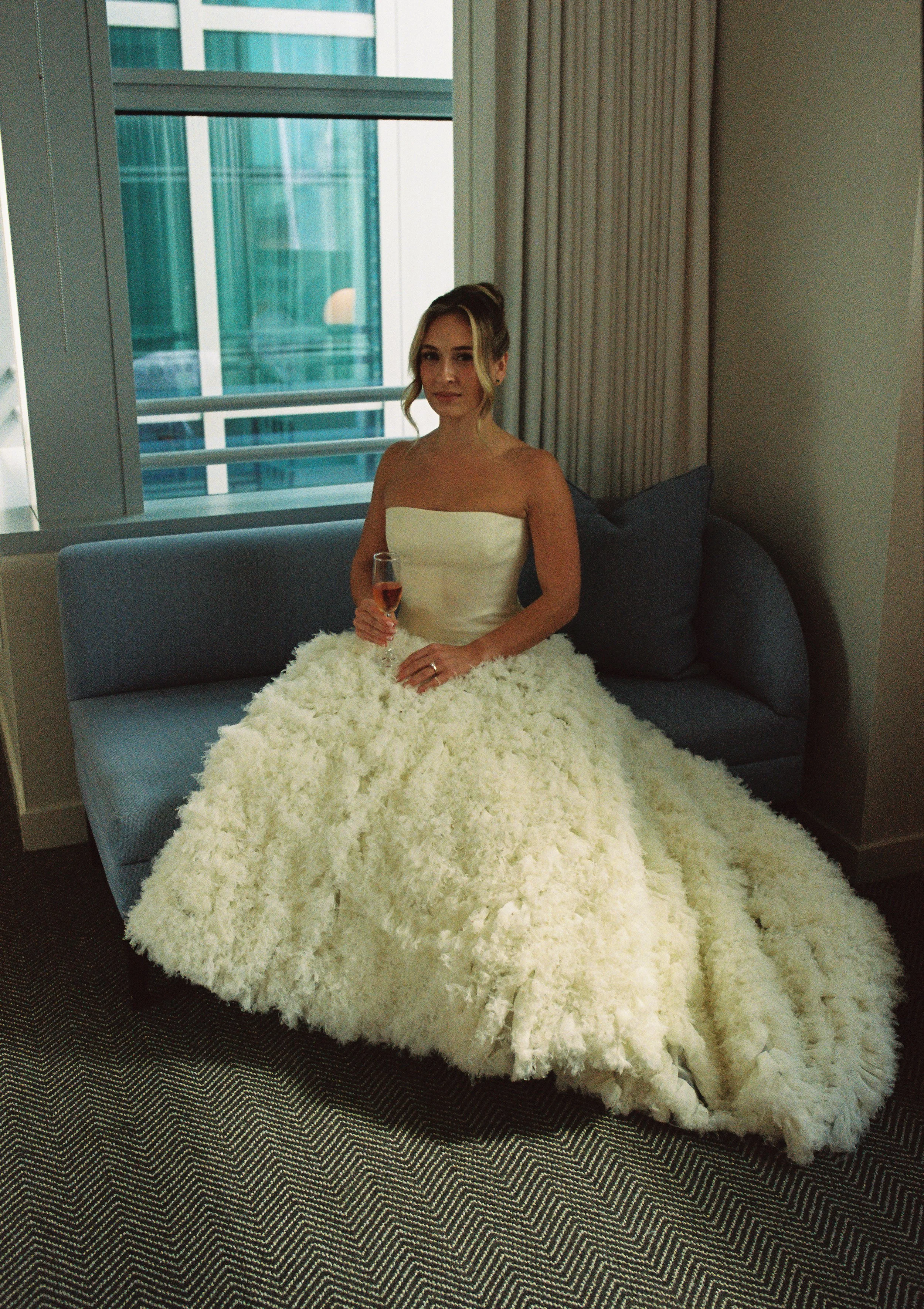 Bride sitting on a modern sofa holding a champagne glass while wearing a textured ballgown wedding dress before her ceremony.
