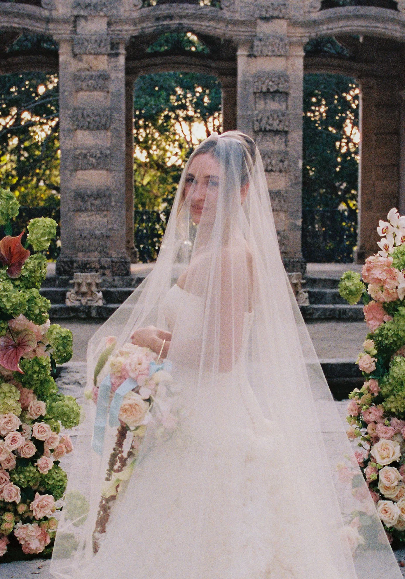 Bride standing beneath a veil between pastel floral arrangements during a romantic Vizcaya Museum and Gardens wedding ceremony.