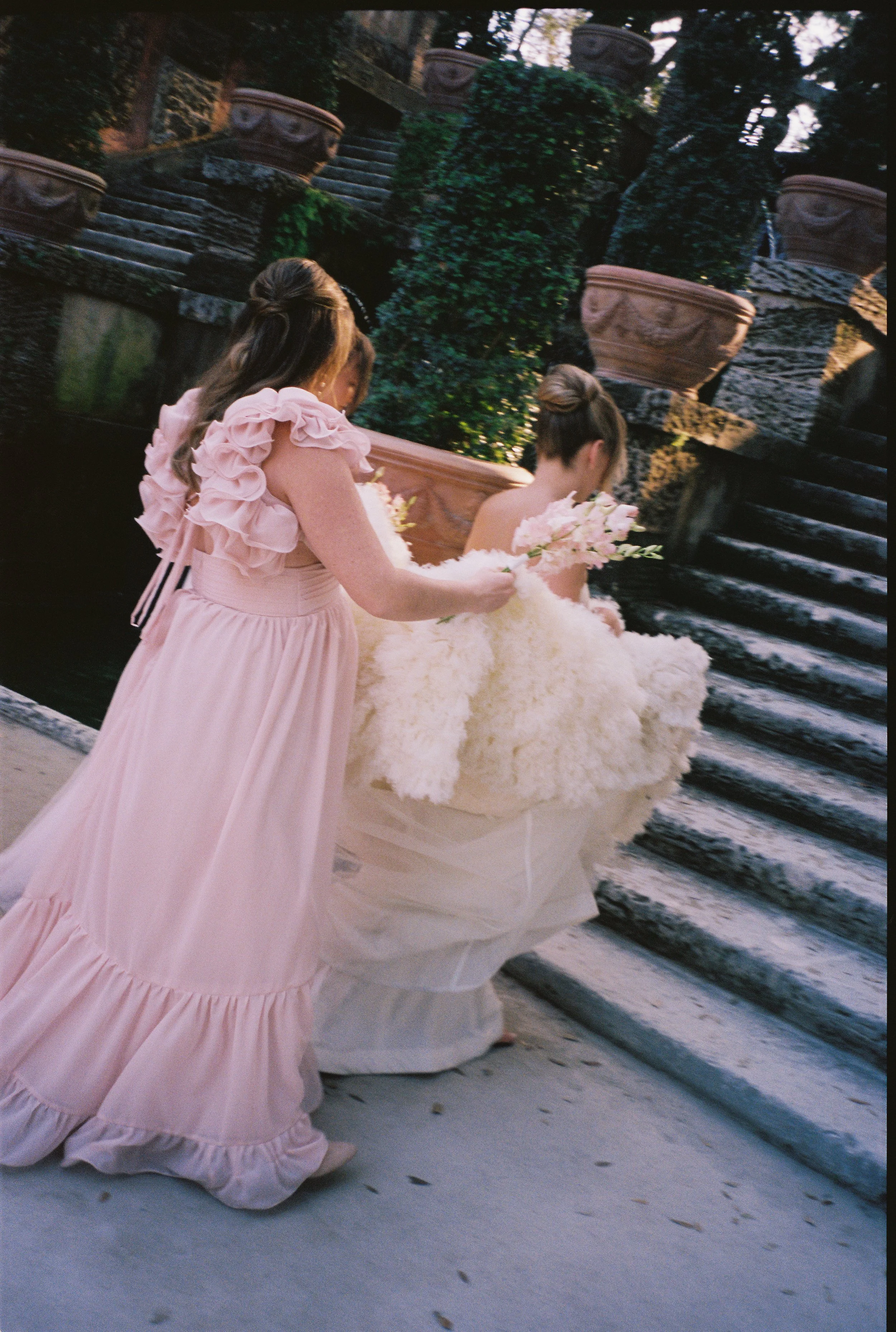 Bridesmaid helping the bride carry her textured wedding gown up stone garden steps at Vizcaya Museum and Gardens.