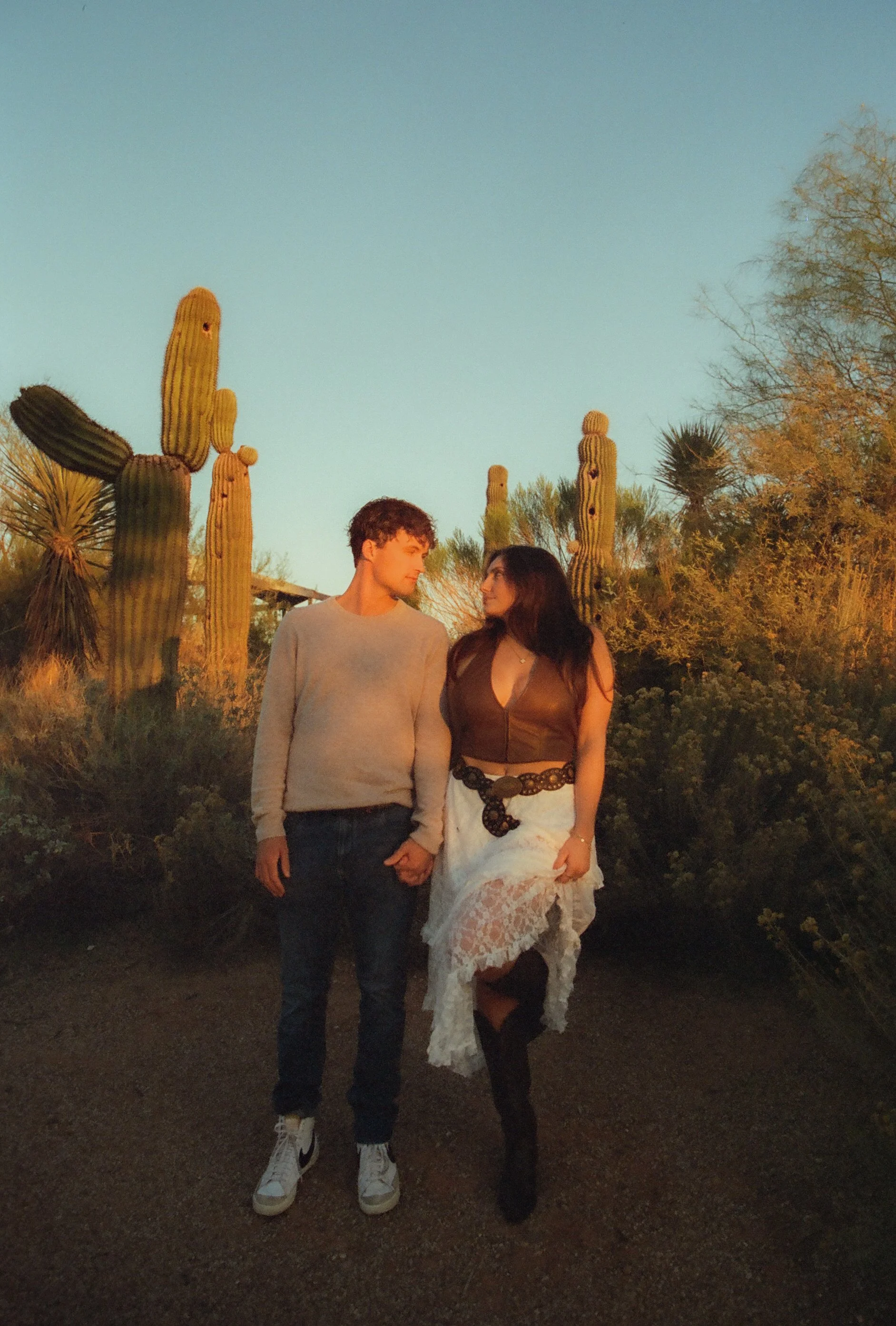 Couple walking hand-in-hand through a sunlit desert path surrounded by saguaro cacti and warm golden light during a couple's photoshoot.