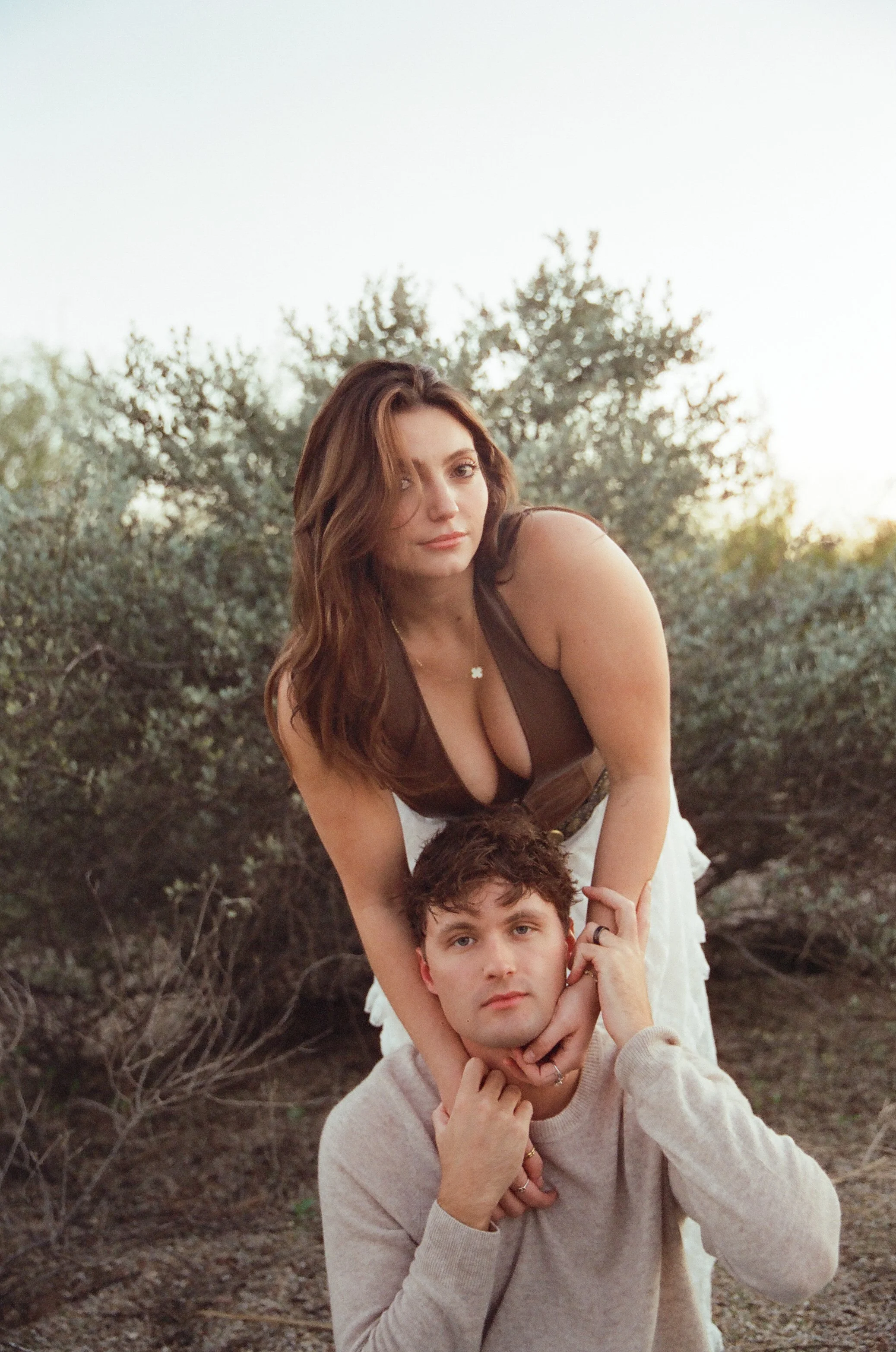Playful portrait of a couple with the woman resting her arms on her partner’s shoulders, framed by soft desert foliage.