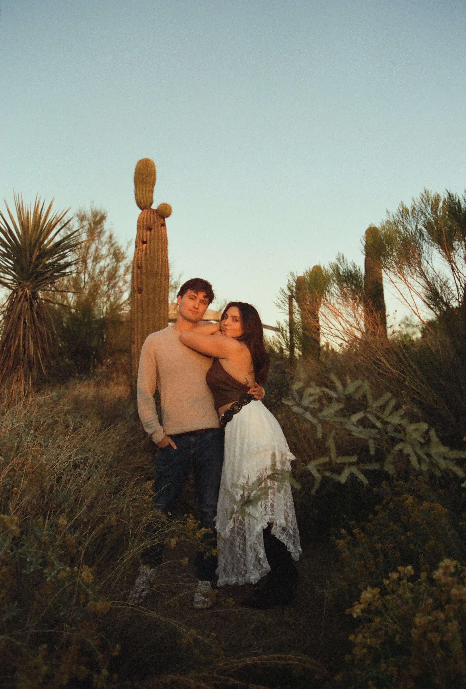 Couple standing close together among desert plants and tall saguaro cacti at golden hour, embracing softly during a couple's photoshoot.
