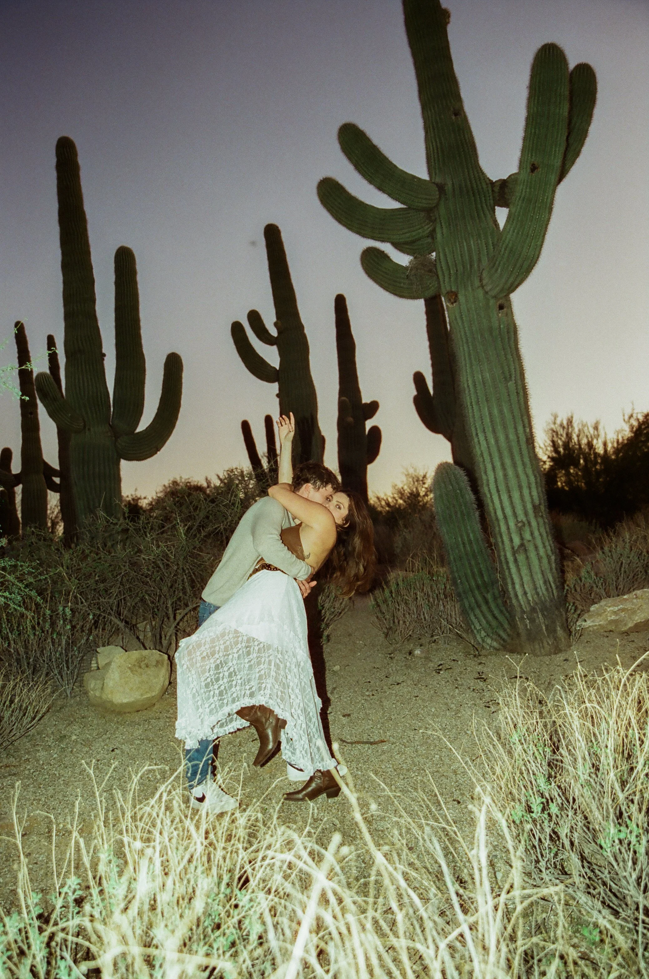 Couple wrapped in each other’s arms in the desert at dusk with dramatic flash lighting and towering saguaro cacti in the background during a couple's photoshoot.