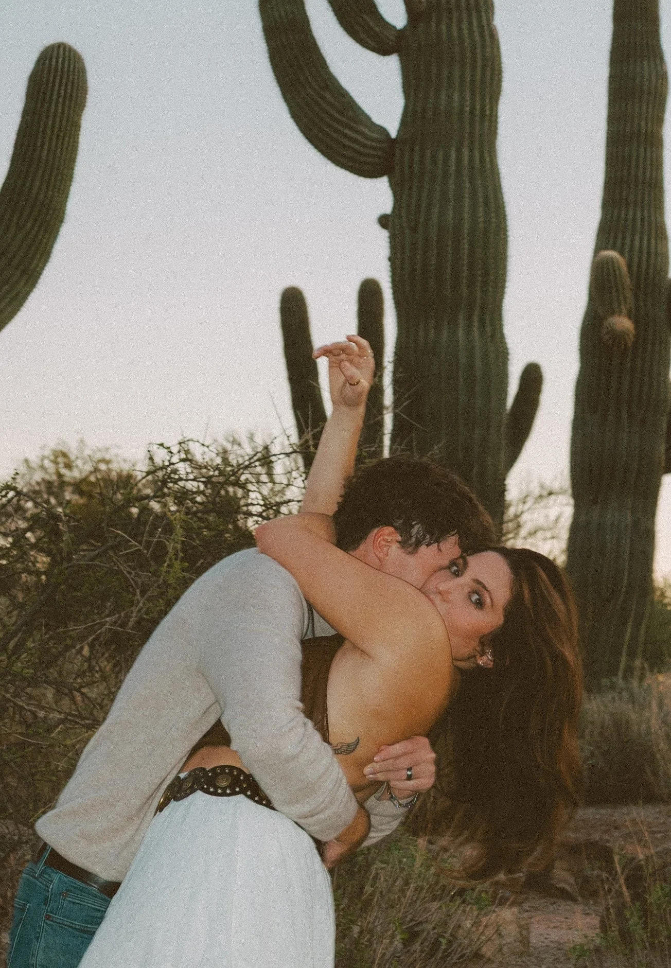Playful, intimate dip pose as the couple embraces among tall saguaro cacti, with warm desert tones and a candid, cinematic feel.