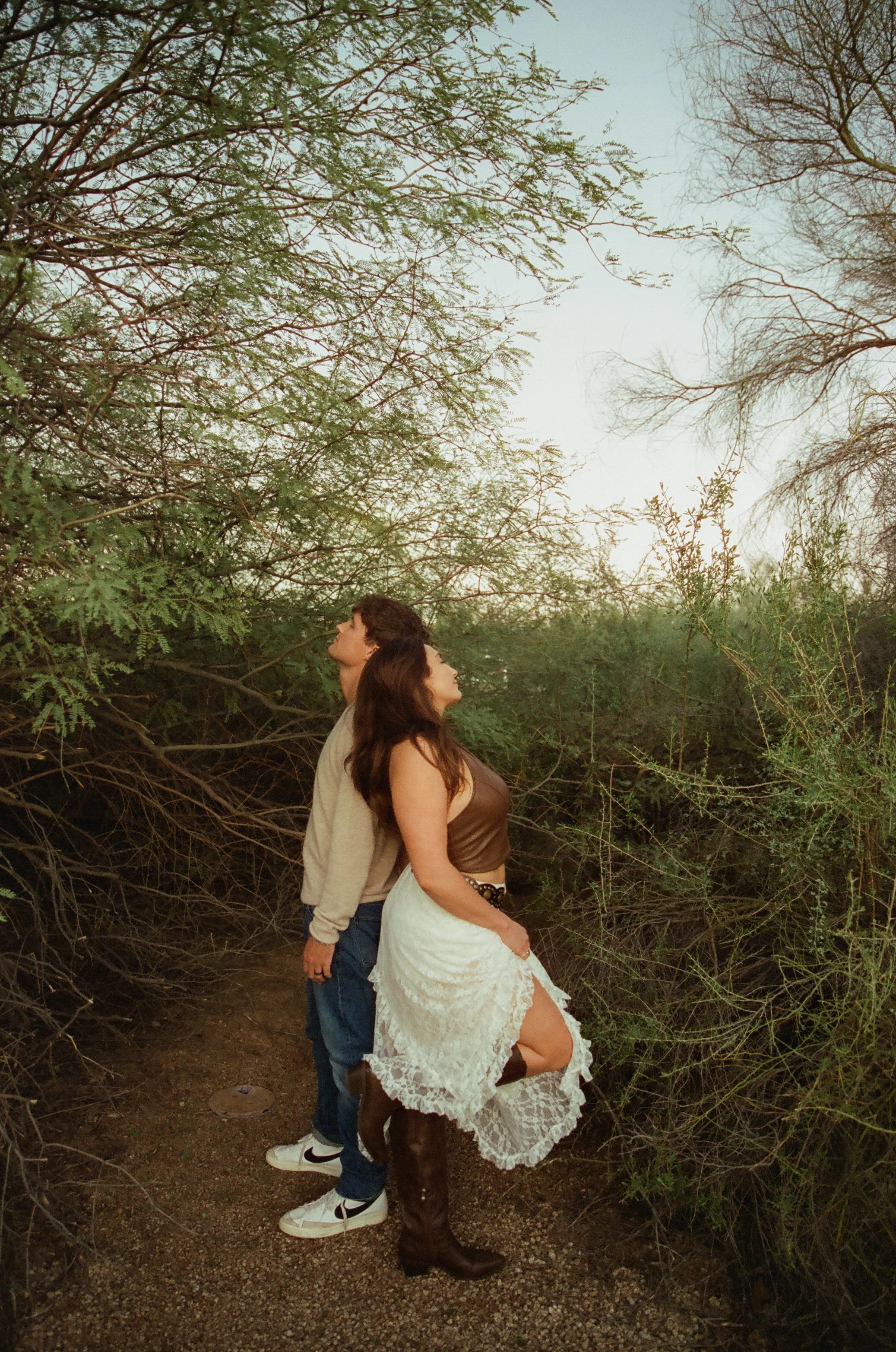 Couple standing back-to-back beneath soft desert greenery, heads tilted upward in a quiet, reflective moment during a couple's photoshoot.