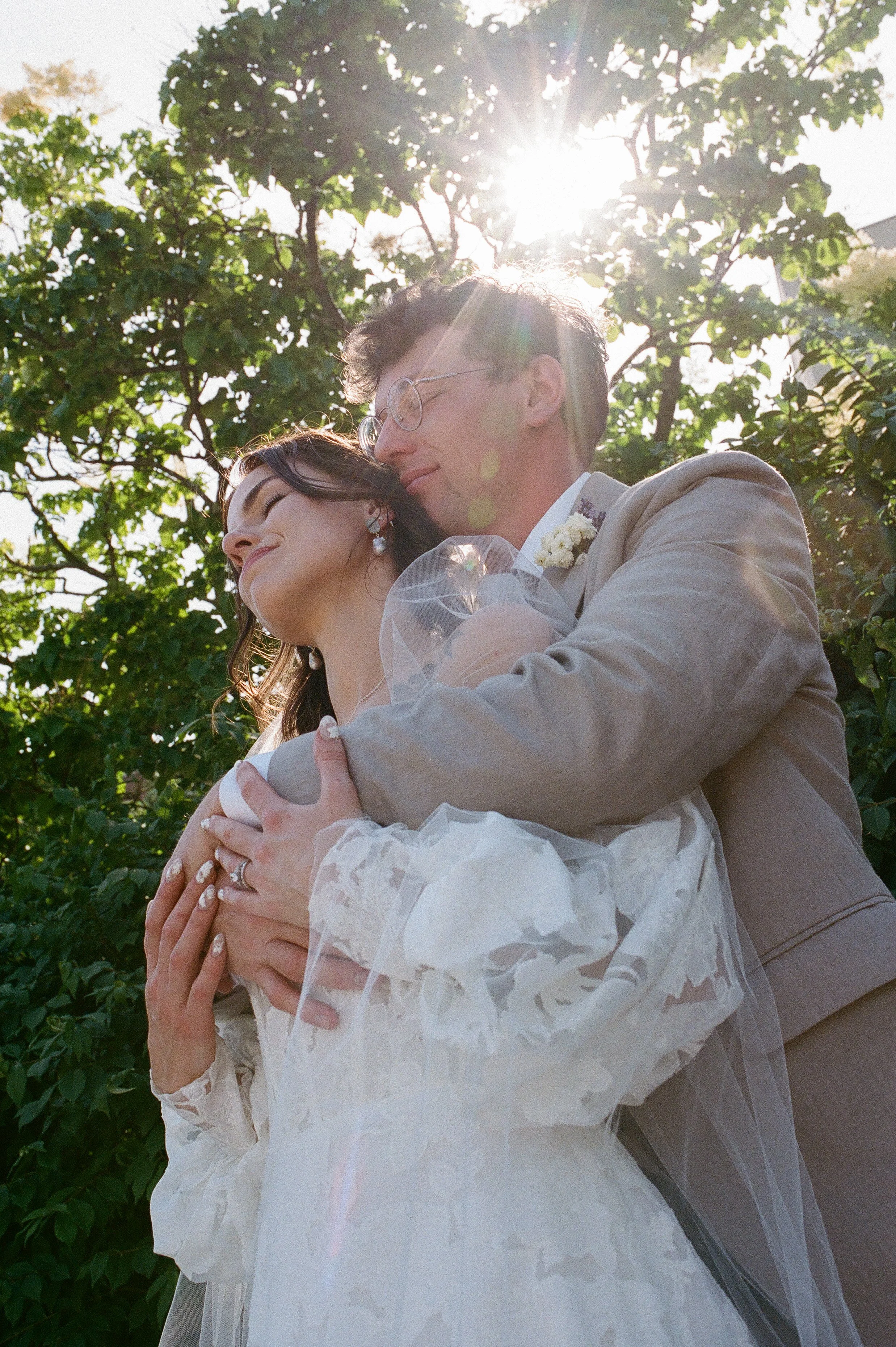 Groom wrapping his arms around the bride under sunlight filtering through trees in a romantic film wedding photography portrait