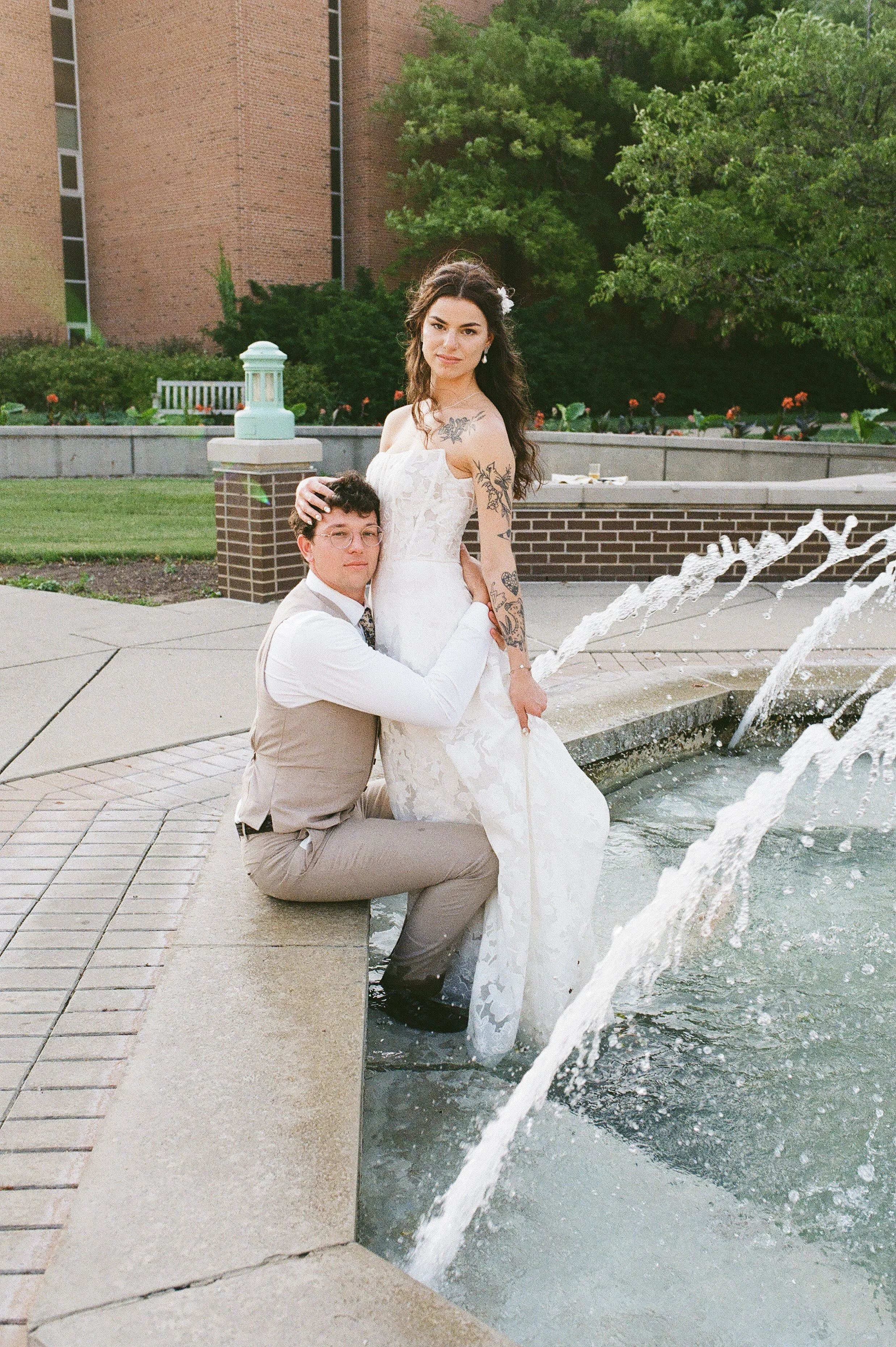Bride standing by a fountain while groom sits beside her, posing together in a modern outdoor setting