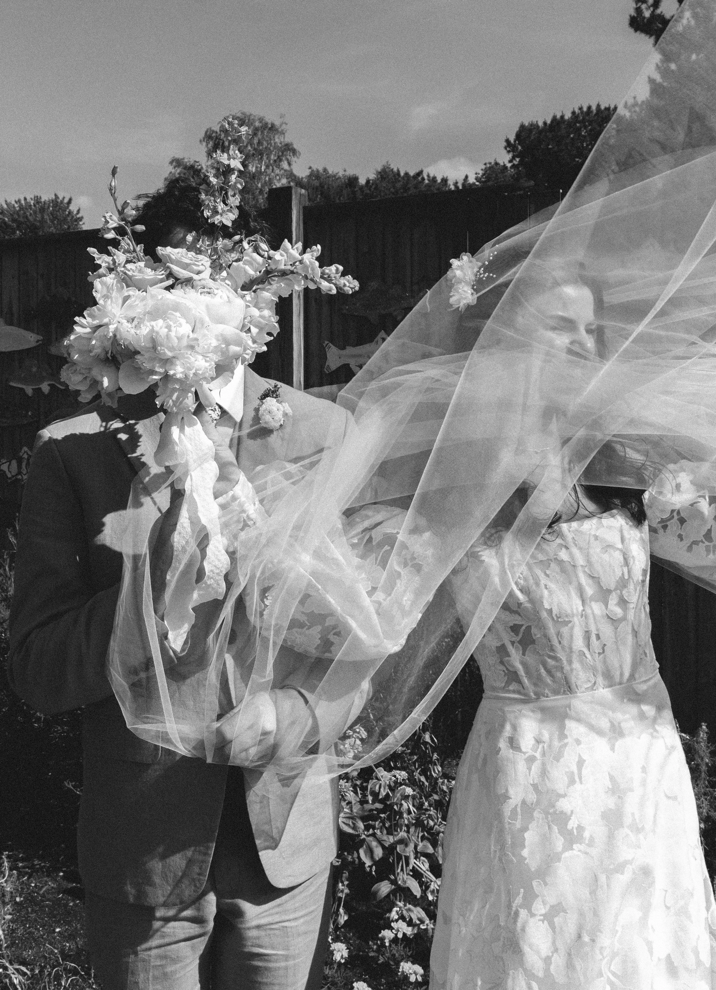 Black and white photo of bride and groom with veil flowing dramatically as they hold a bouquet together