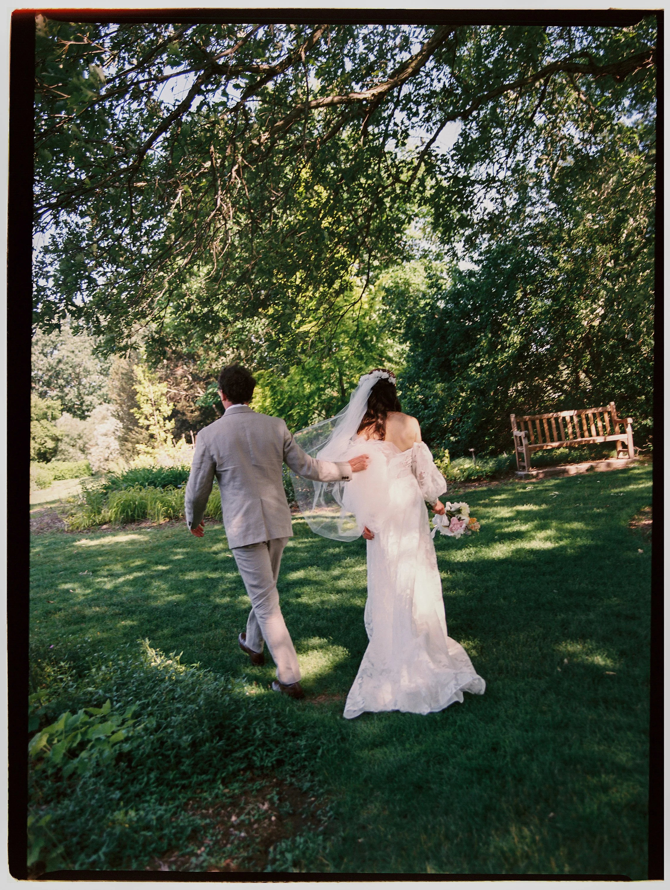 Bride and groom walking hand in hand through a lush garden, captured from behind in a romantic film wedding photography moment