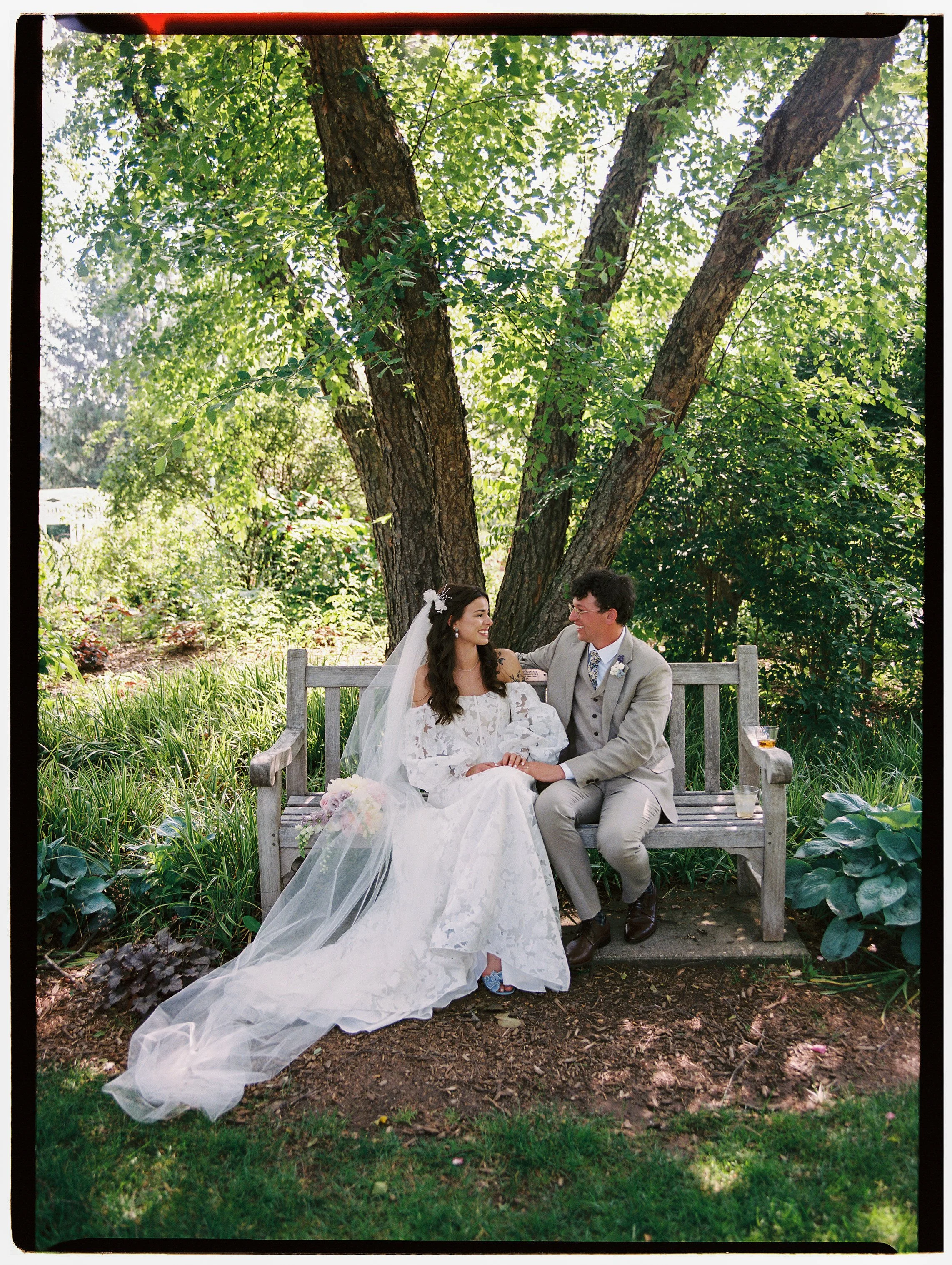 Bride and groom sitting together on a wooden bench under a large tree, sharing a quiet, intimate moment