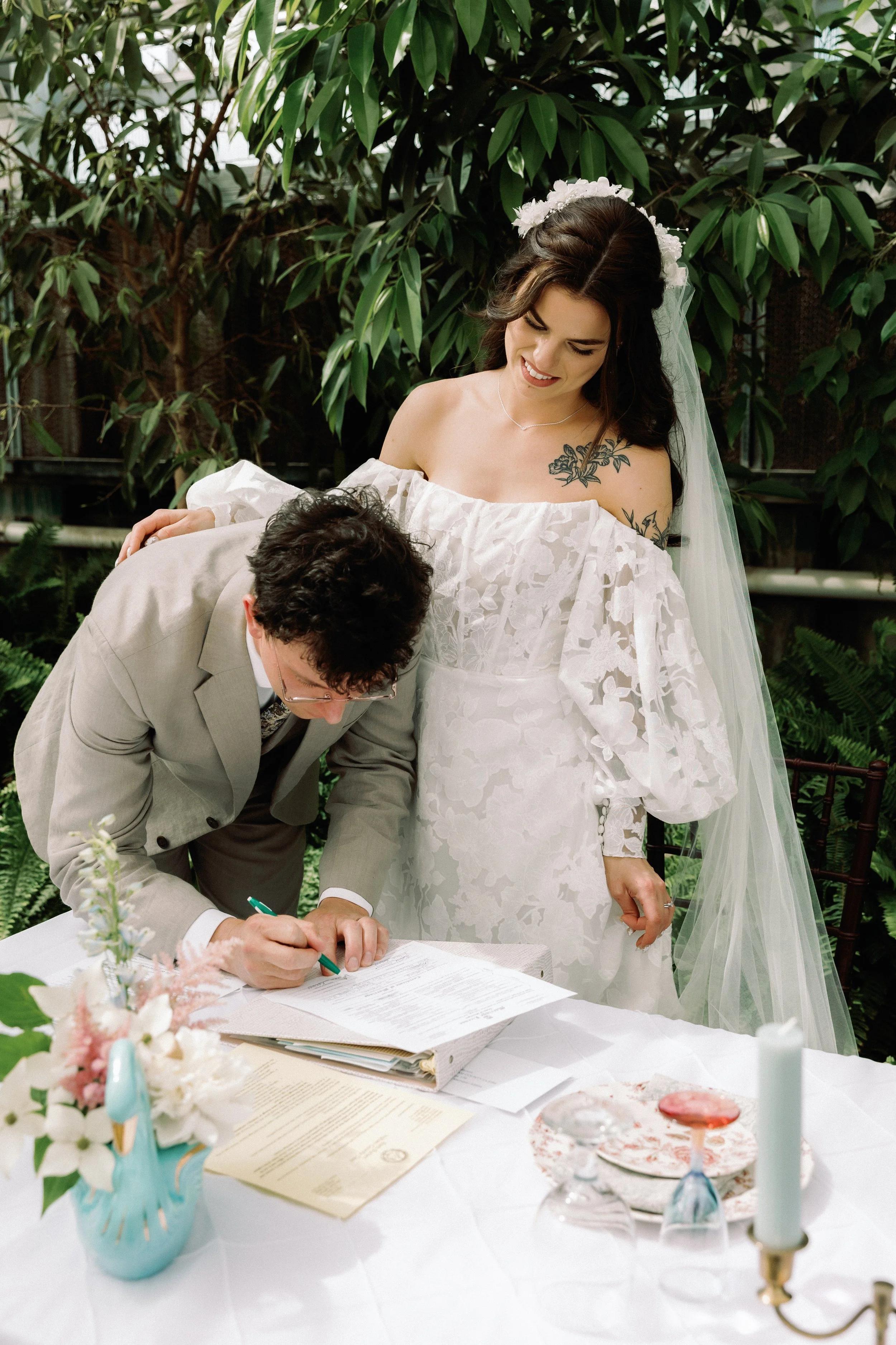 Groom signing the marriage license while bride stands beside him in a greenhouse setting