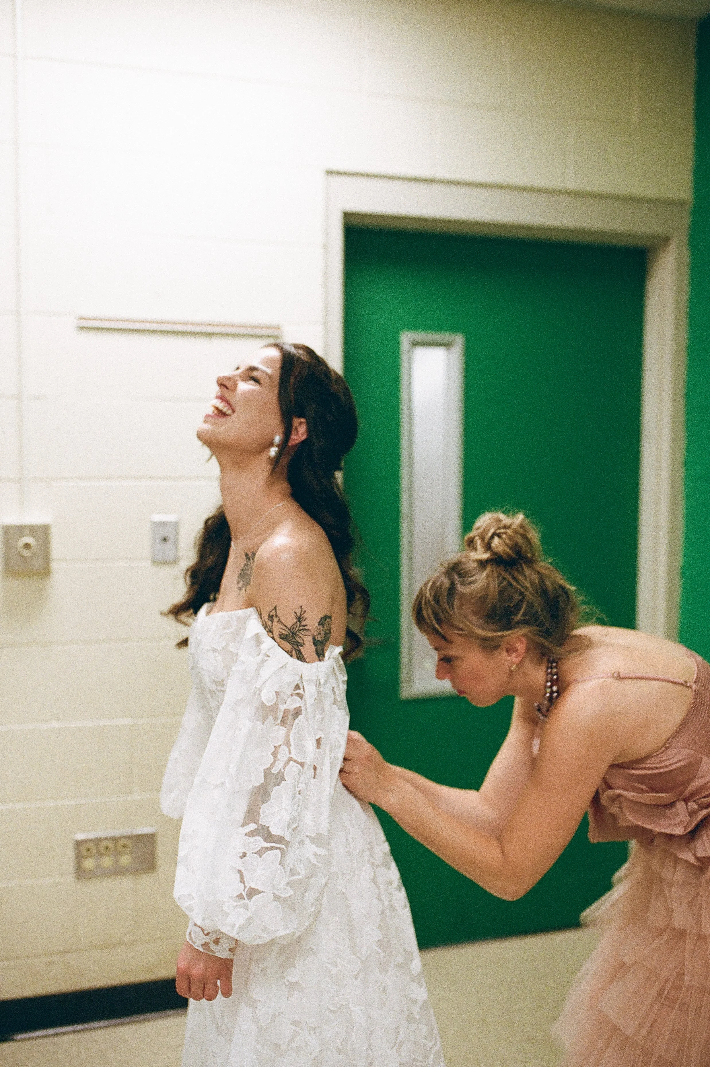 Bride laughing while a bridesmaid buttons the back of her lace wedding dress in a candid getting ready moment