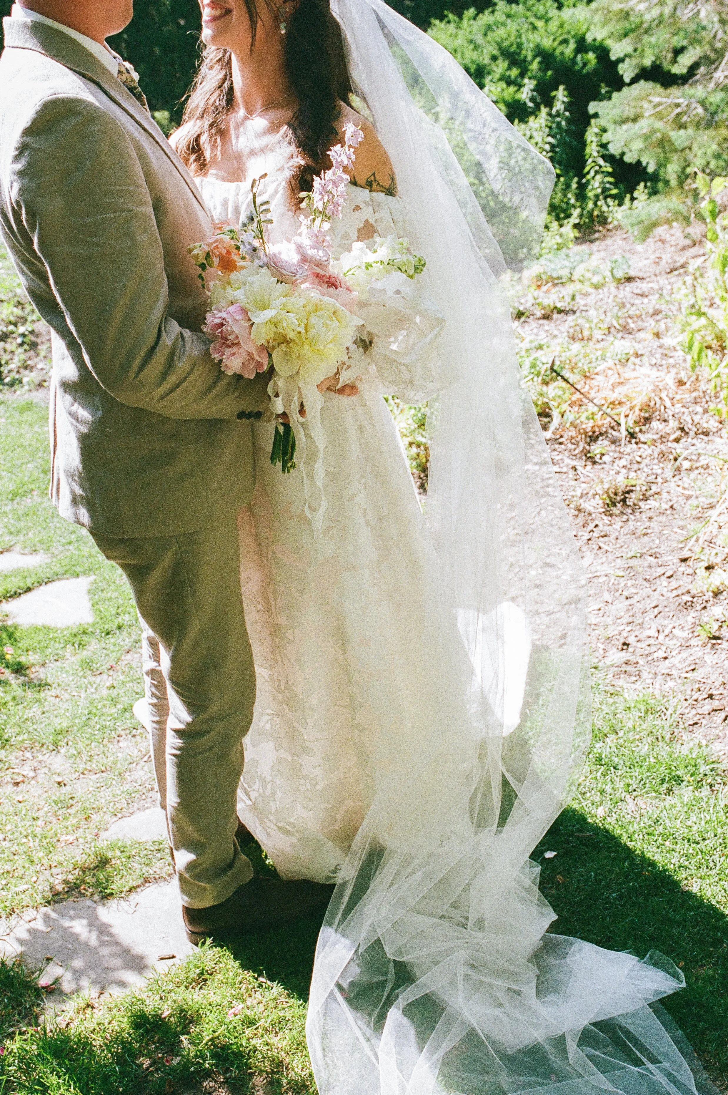 Bride and groom standing close together outdoors, holding a soft pastel bouquet with her veil flowing behind her in a romantic film wedding photography moment