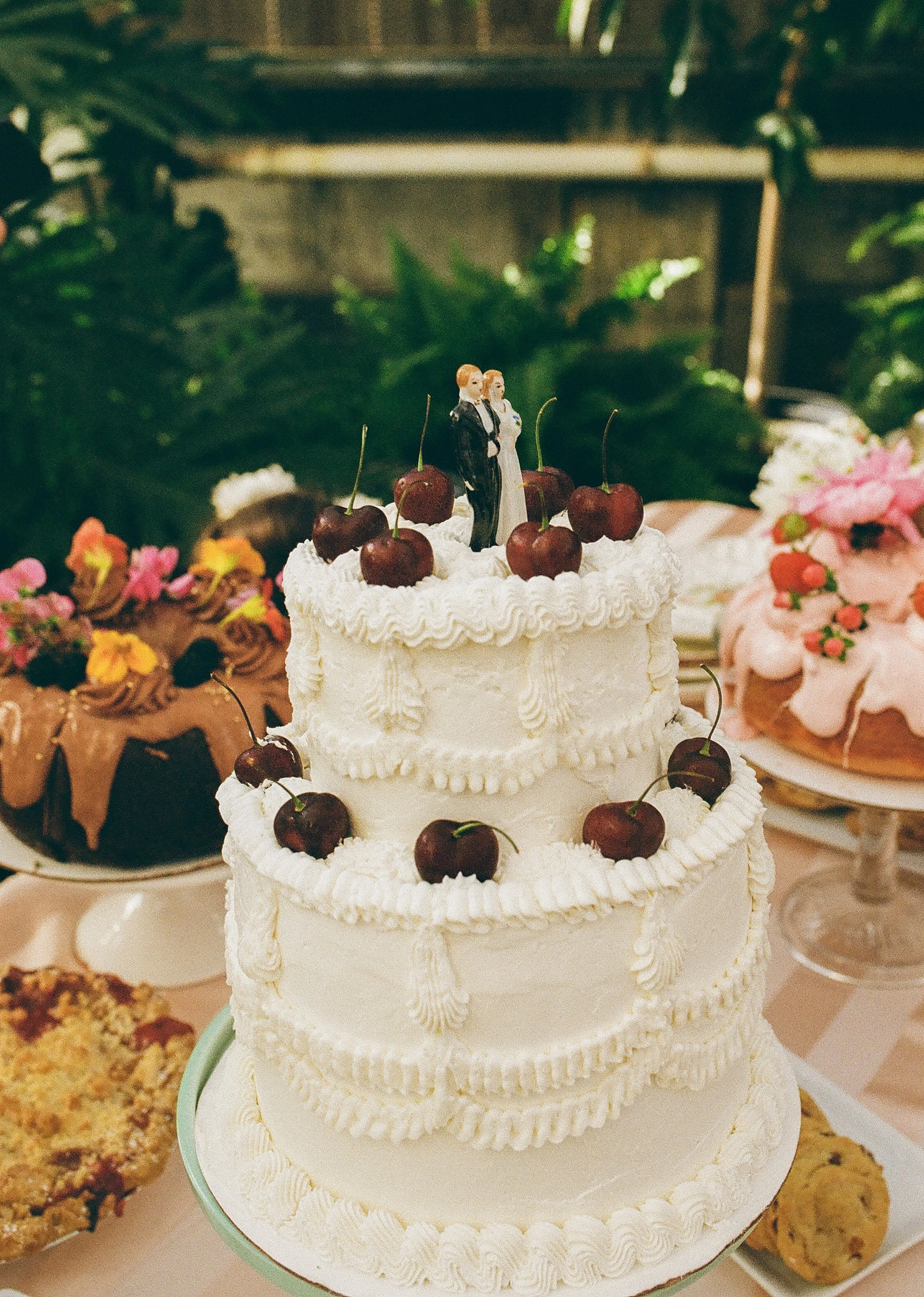 Two-tier wedding cake topped with cherries and a bride and groom figurine, styled among desserts in a film wedding photography aesthetic
