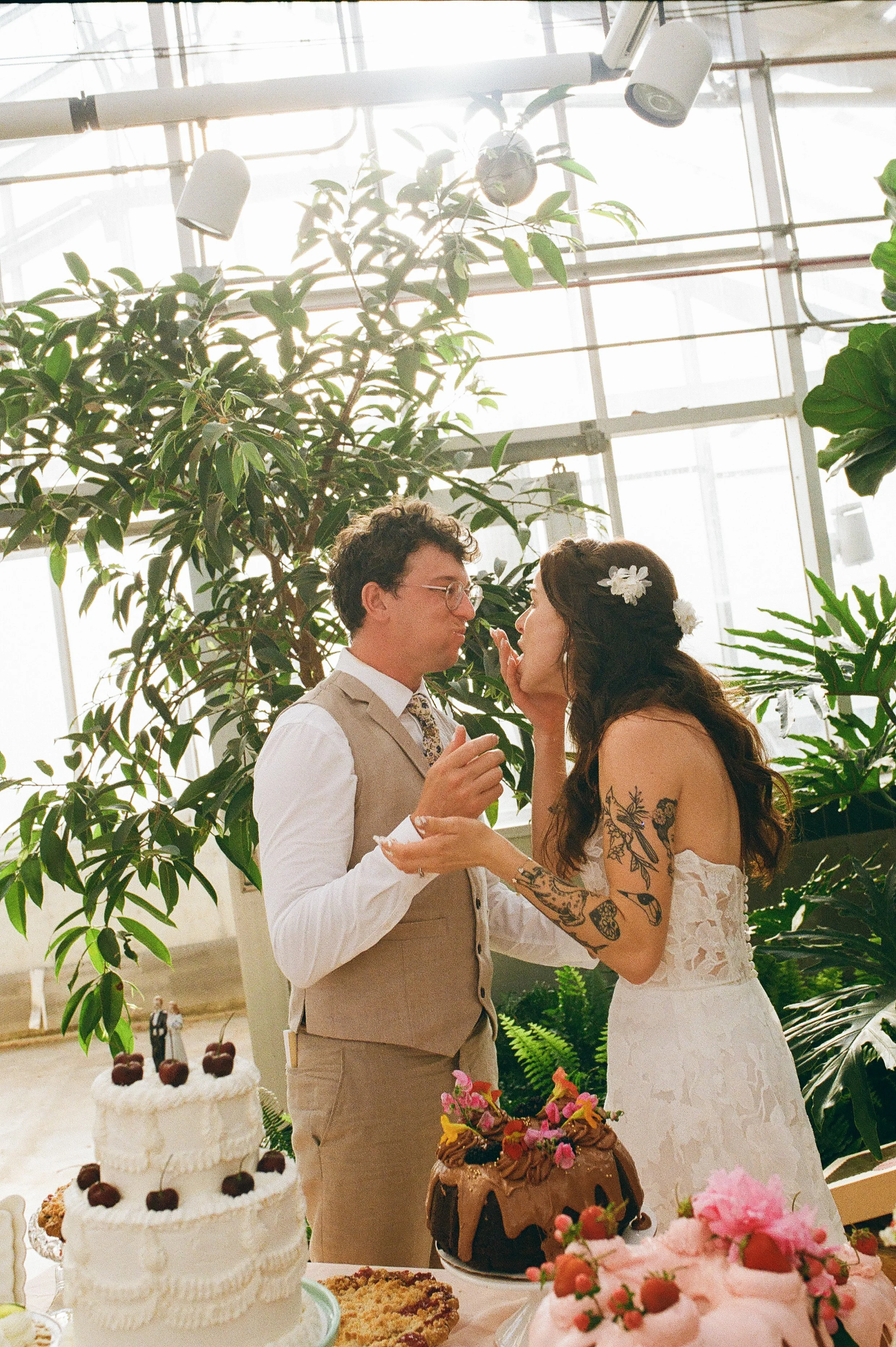Bride and groom feeding each other cake inside a greenhouse reception filled with natural light and greenery