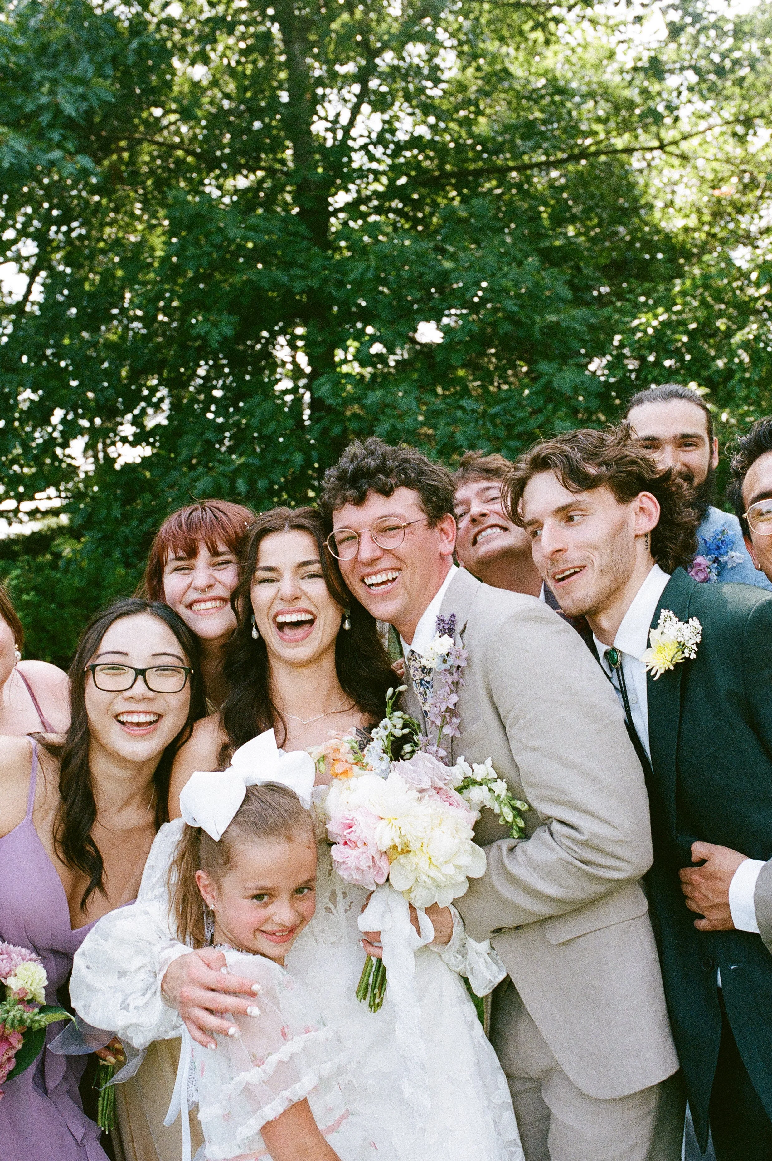 Joyful group portrait of bride and groom surrounded by friends and family, all smiling closely together outdoors