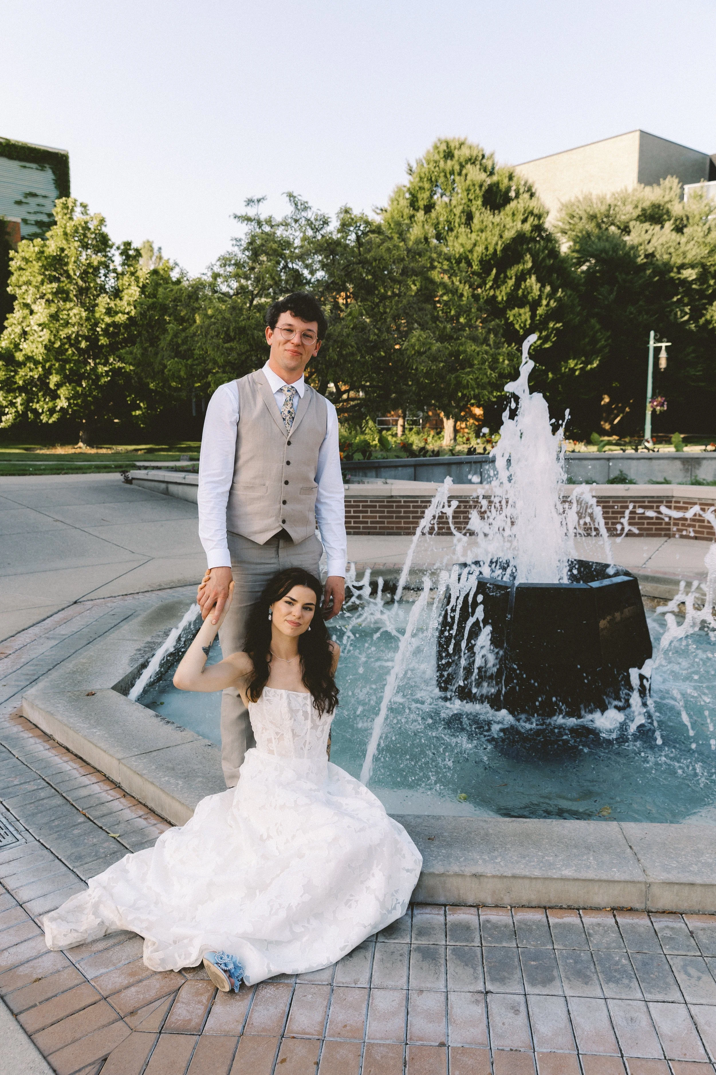 Bride sitting on the edge of a fountain holding the groom’s hand while he stands behind her, surrounded by greenery in a romantic film wedding photography style