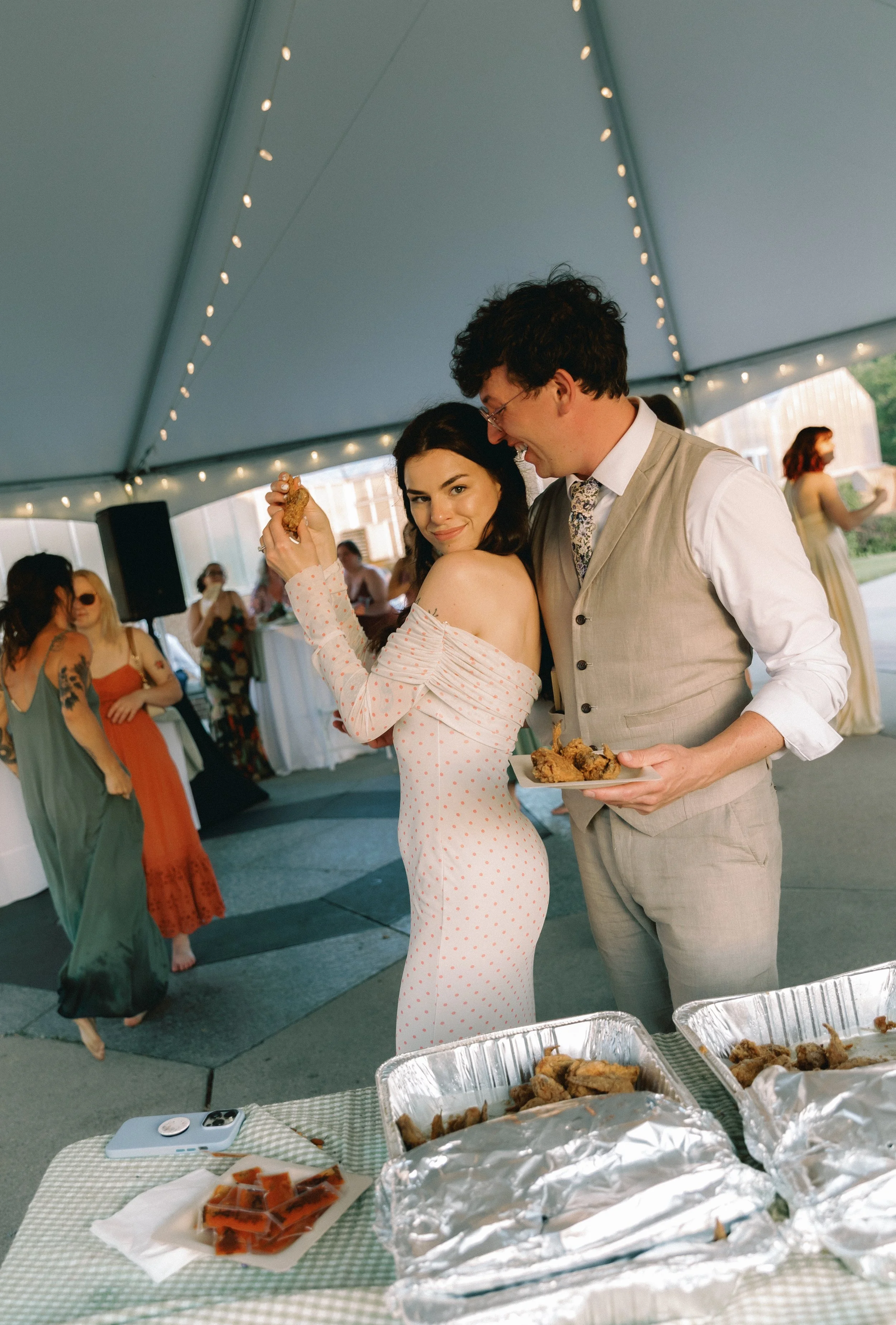 Bride in a fitted polka dot dress holding fried chicken while standing close to the groom under a reception tent, captured in a candid, playful moment