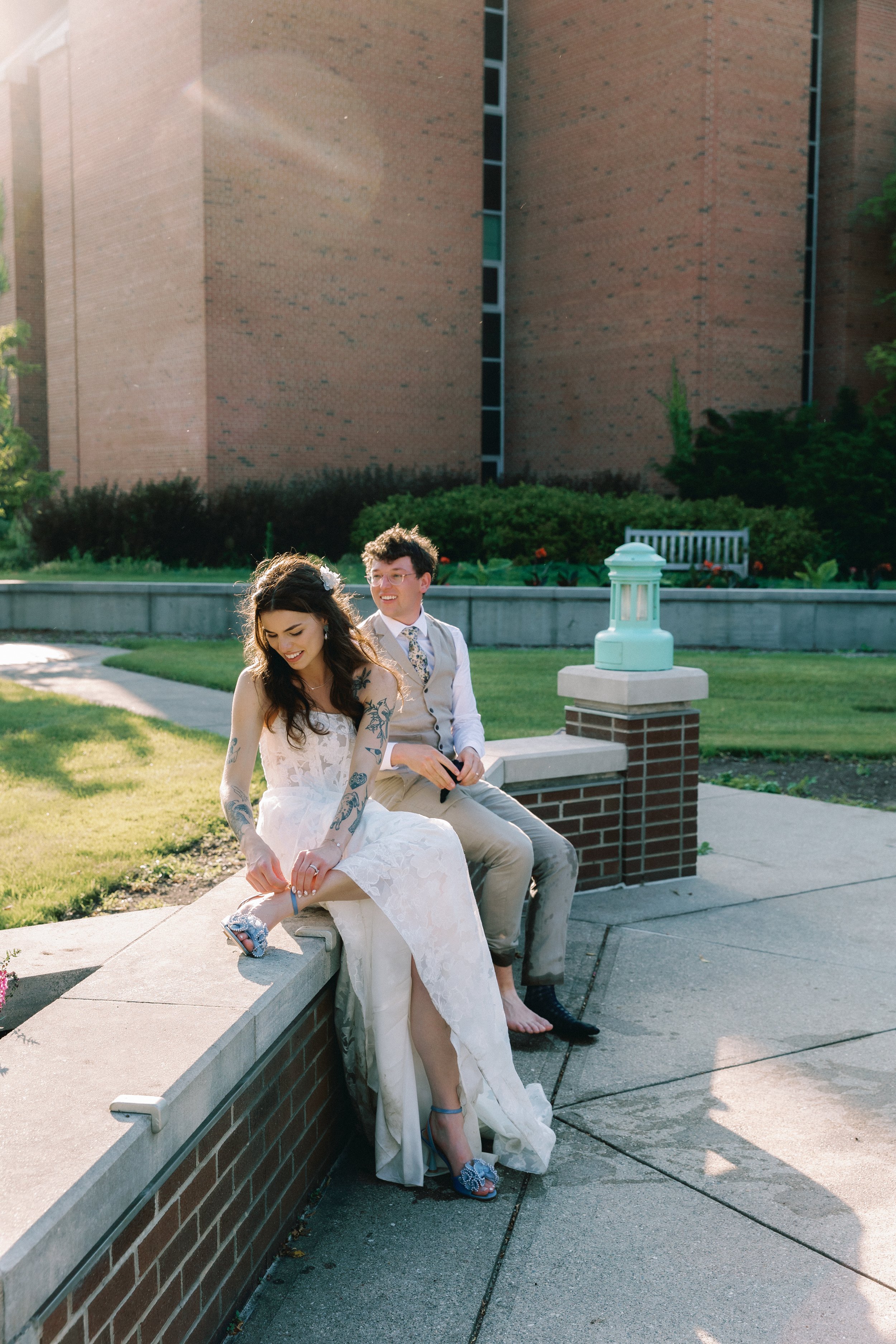 Bride and groom sitting on the ledge, the bride looking down at her bridal bouquet, while her groom is behind her looking into the distance.